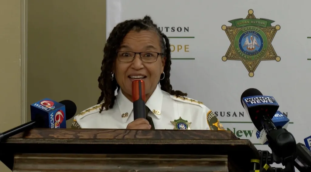 Sheriff Susan Hutson speaking at a podium with news microphones.