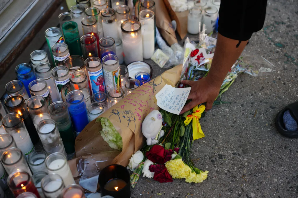 A person's hand places flowers, a stuffed animal, and a handwritten note at a makeshift memorial of lit candles and flowers.