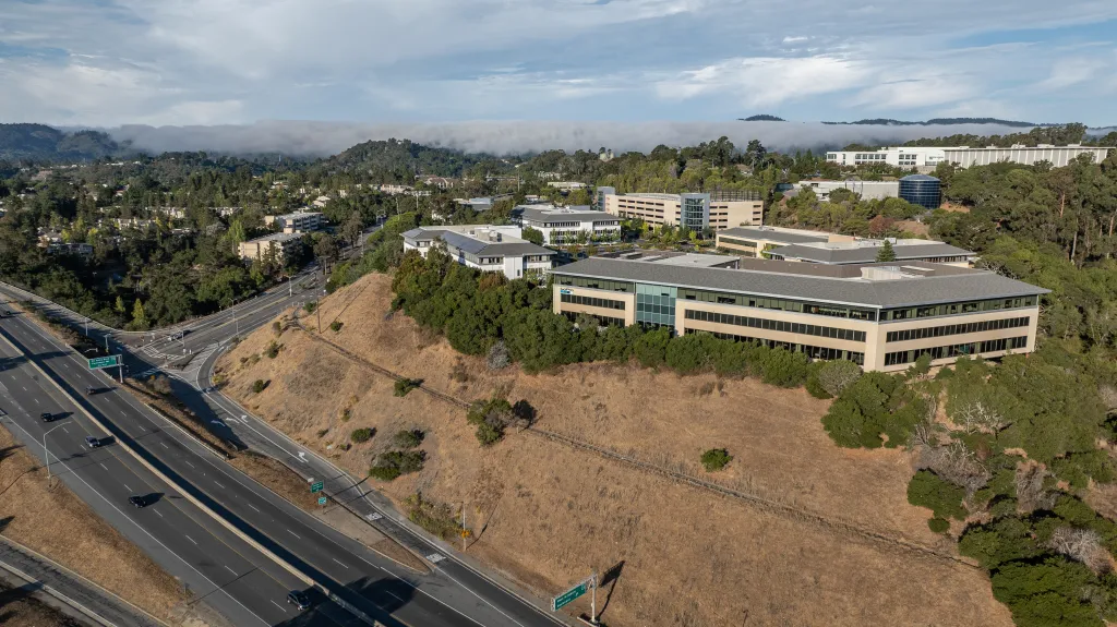 Aerial drone view of the GoPro headquarters at 3025 Clearview Way in San Mateo, California, with a highway in the foreground and hills partially covered by fog in the background.