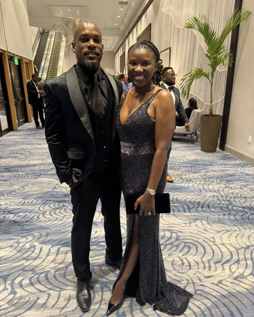 A man in a black tuxedo and a woman in a sparkling black gown posing in a hallway.