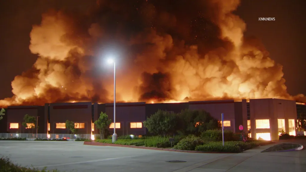 A large warehouse engulfed in flames at night, with thick smoke filling the sky.