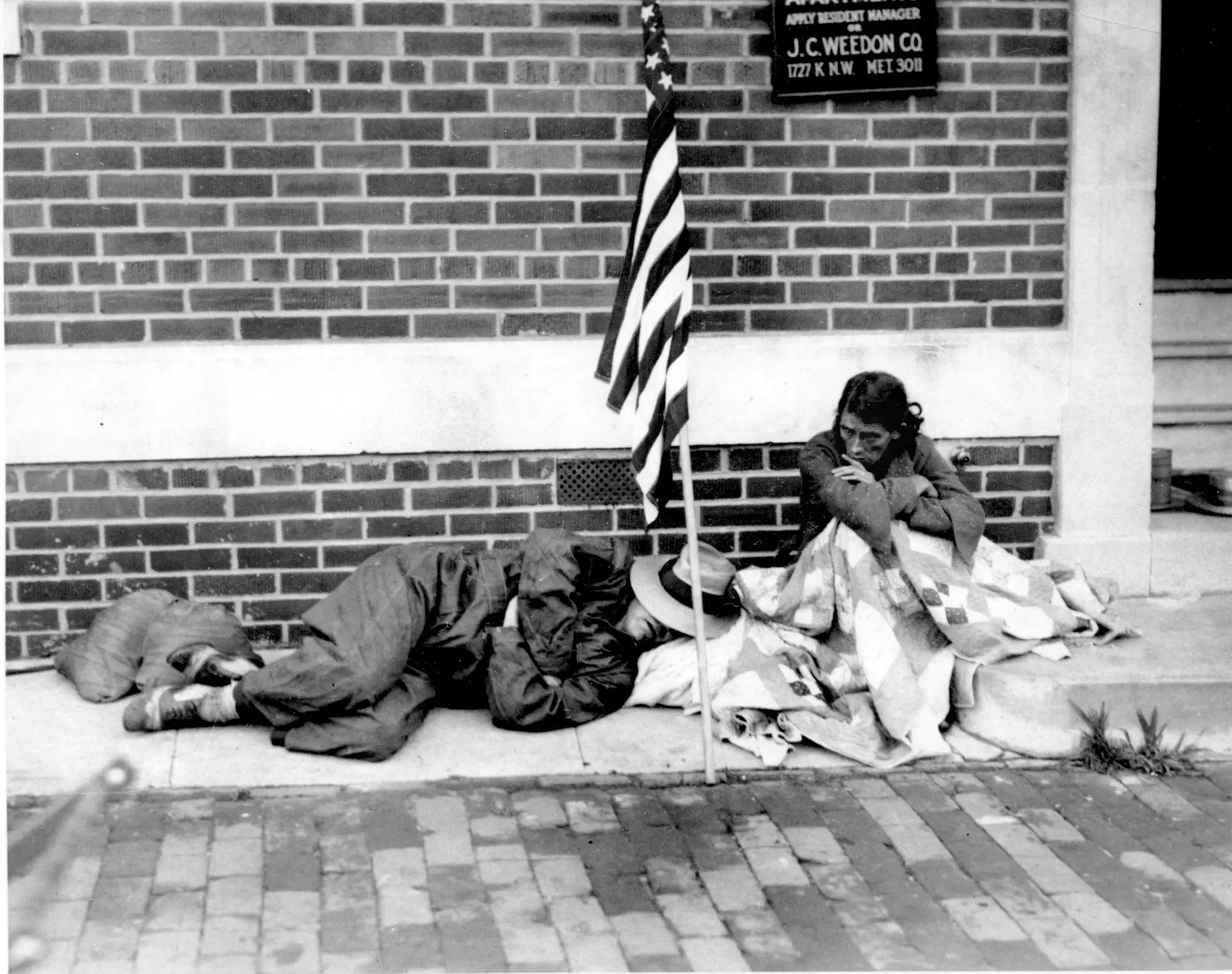 A homeless veteran sleeps on the sidewalk during the Great Depression as his wife sits wrapped in blankets.