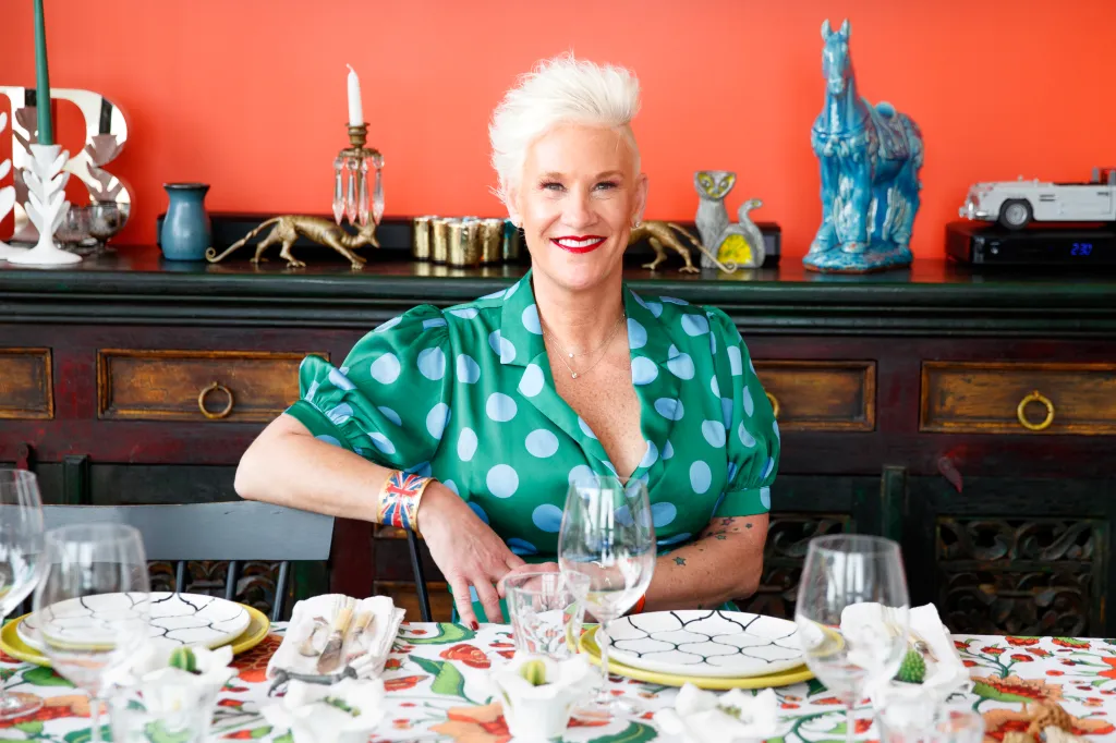 Chef Anne Burrell smiling while seated at a dining table with a colorful tablecloth and place settings.