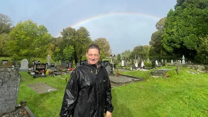 A smiling man in a black raincoat stands in a cemetery with a rainbow overhead.