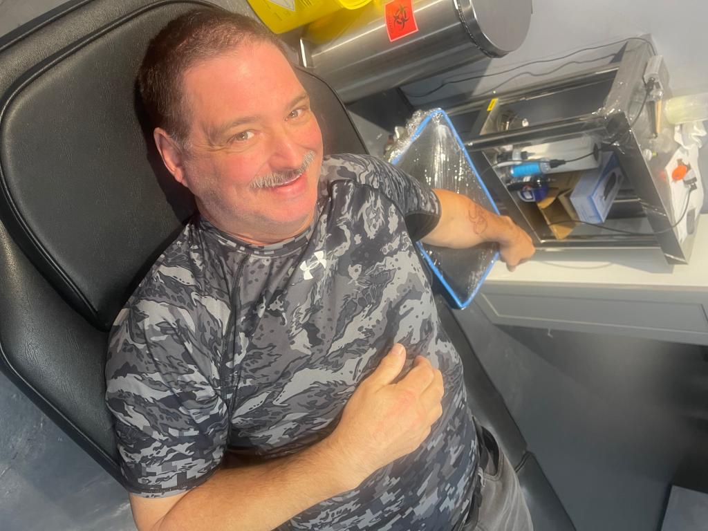 A 61-year-old Costco employee, Randy Corrigan, smiling while lying in a chair, likely during a tattooing session, with a visible arm tattoo.