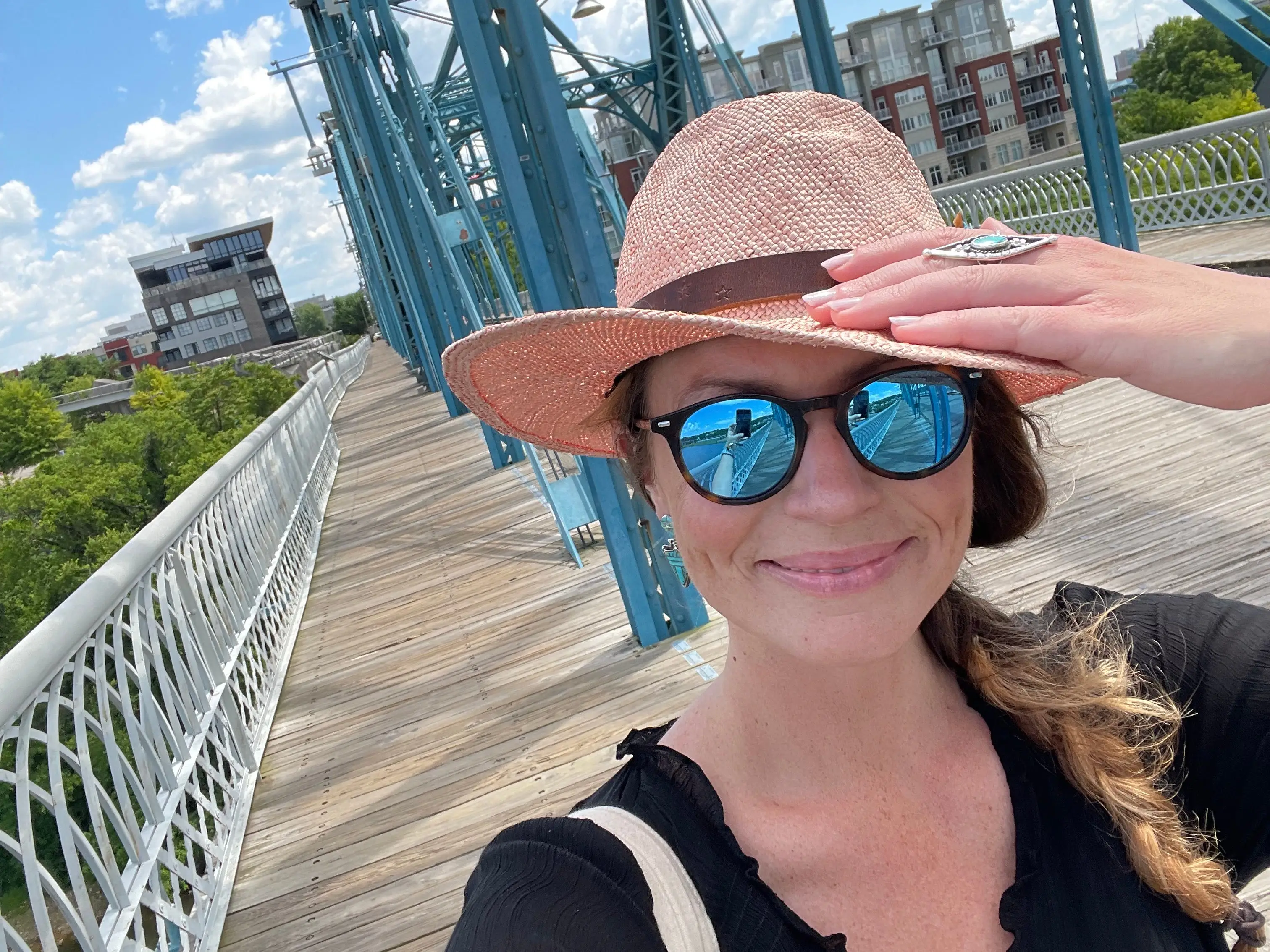 Emily, wearing a black shirt, sunglasses, and a pink hat, takes a selfie on a bridge.