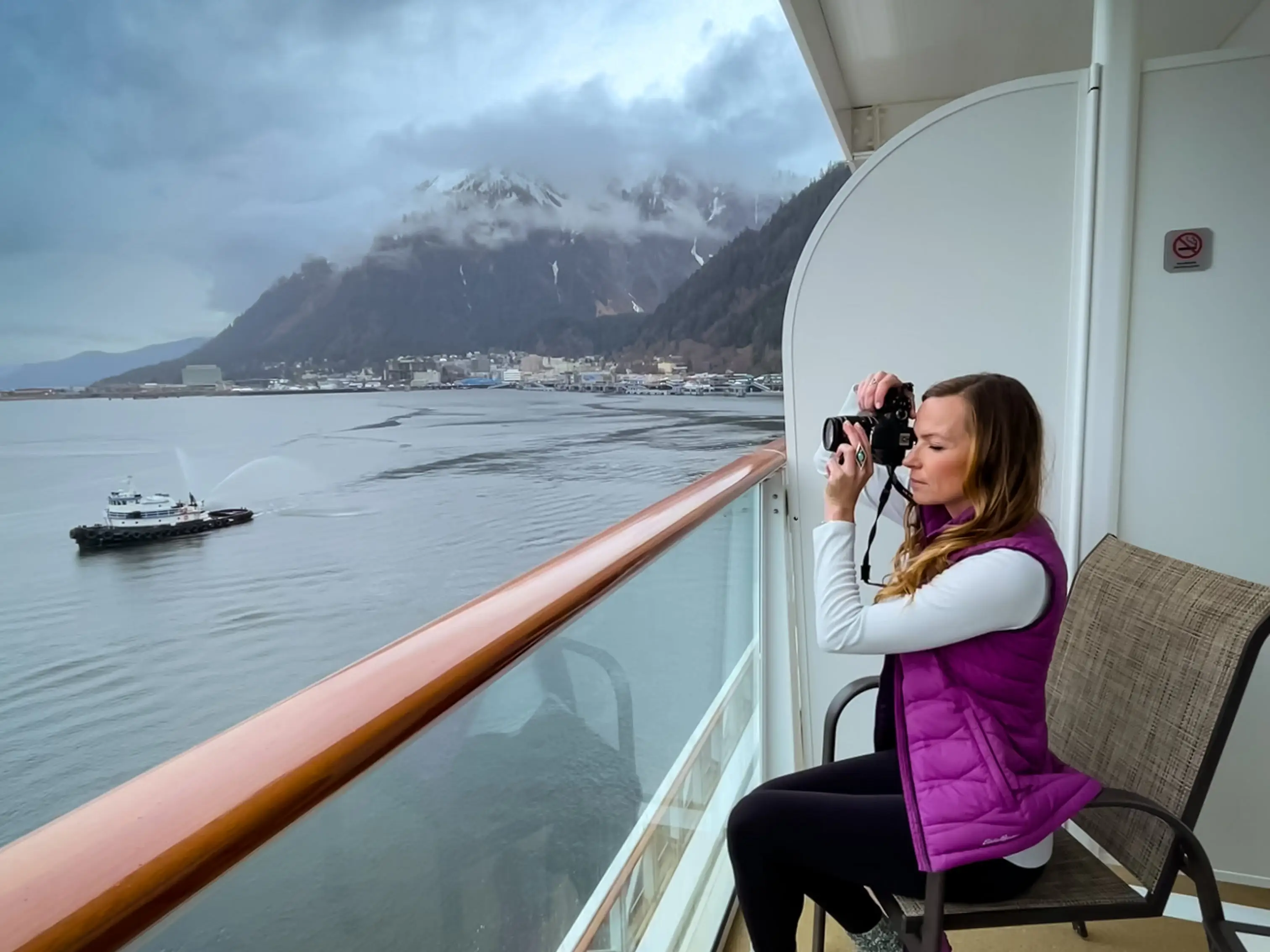 Emily, wearing a purple puffer vest, a white long-sleeve shirt, black pants, and purple sneakers, takes a photo on the balcony of an Alaskan cruise.