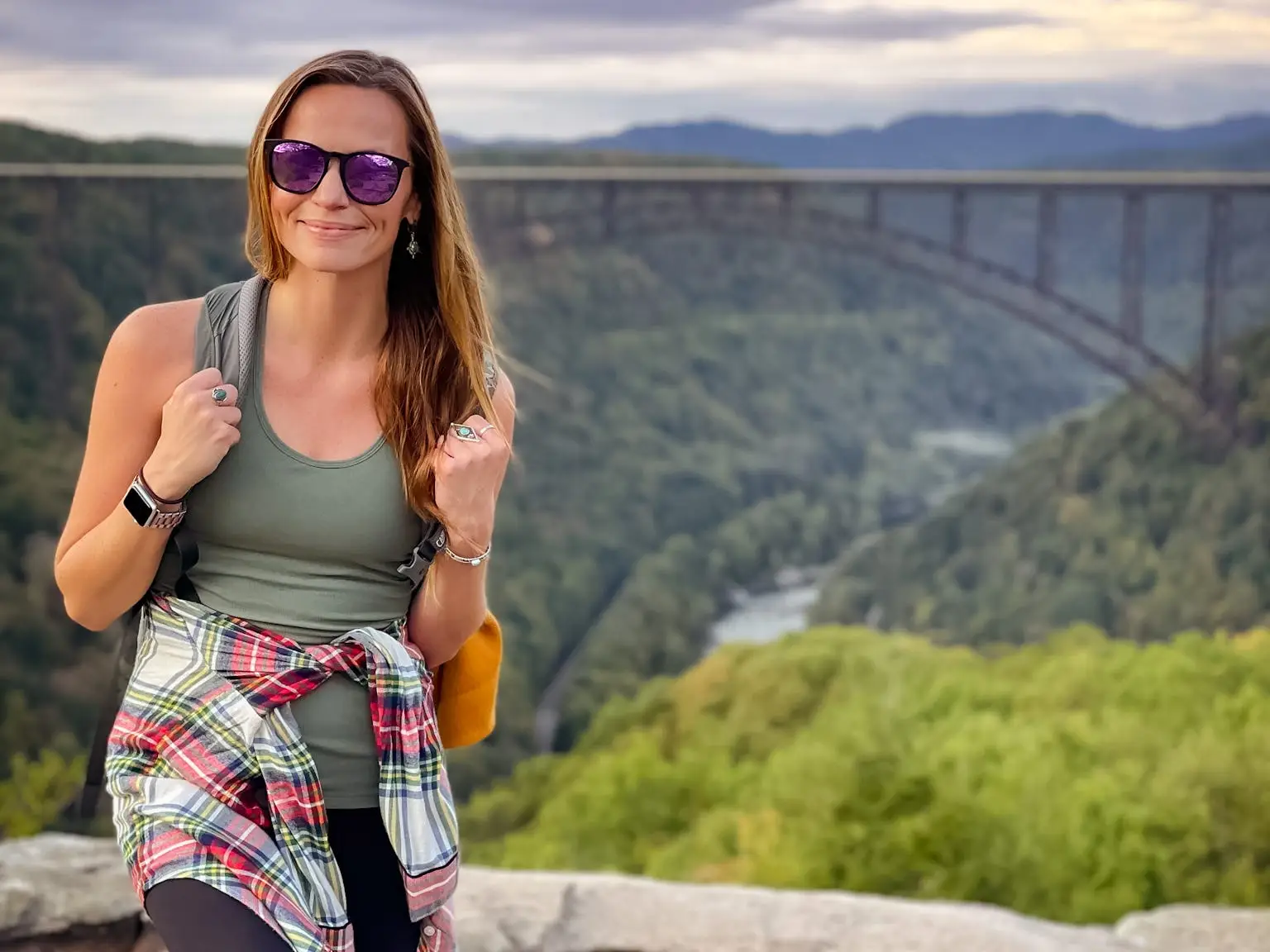 Emily, wearing sunglasses, a green tank top, black leggings, and a flannel around her waist, stands in front of New River Gorge Bridge.