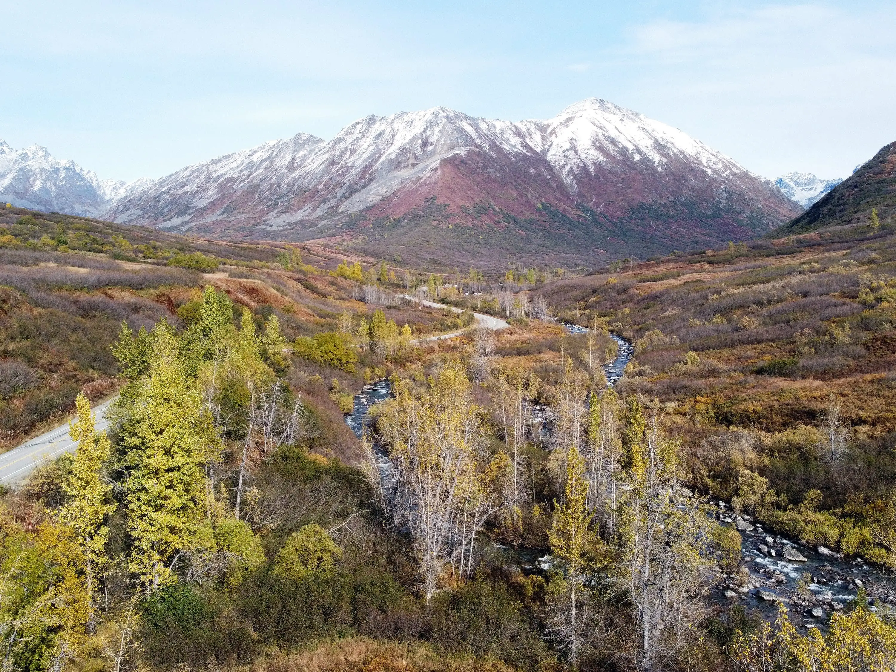 High view of Hatcher Pass Road (left) with a snowcapped mountain (center) and a creek (right) view in Palmer, Alaska.