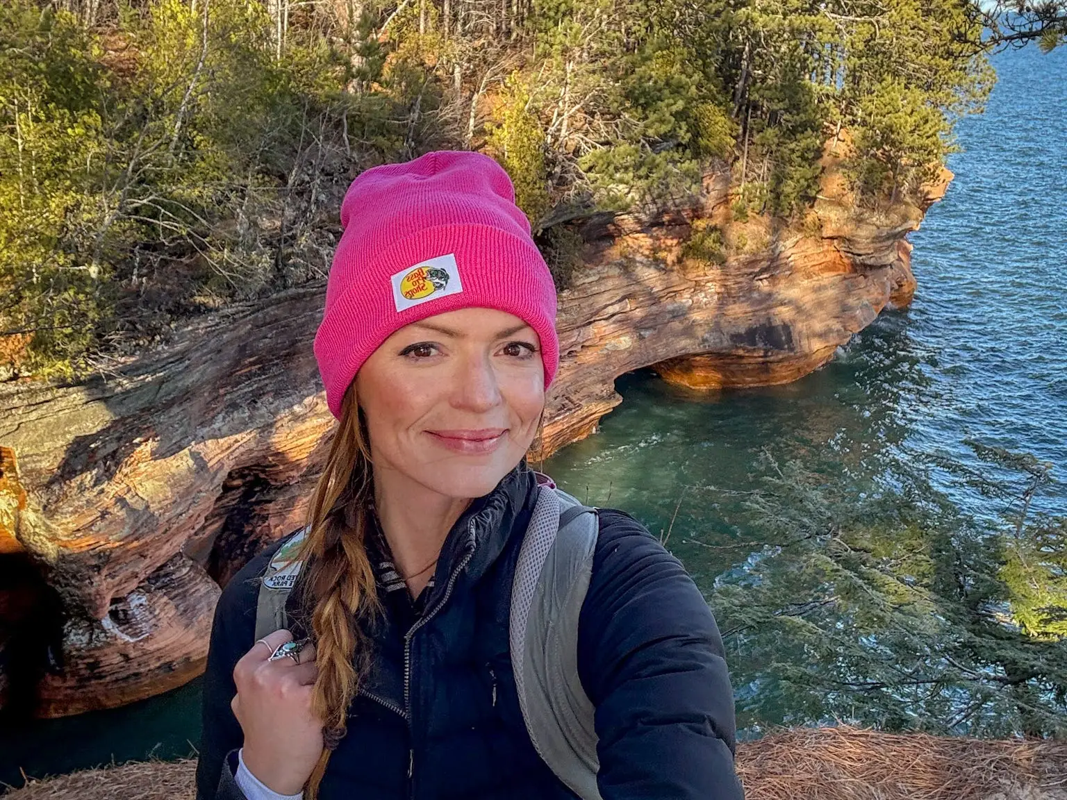 Emily, wearing a backpack, a pink beanie, and a jacket, takes a selfie with trees and a body of water behind her.