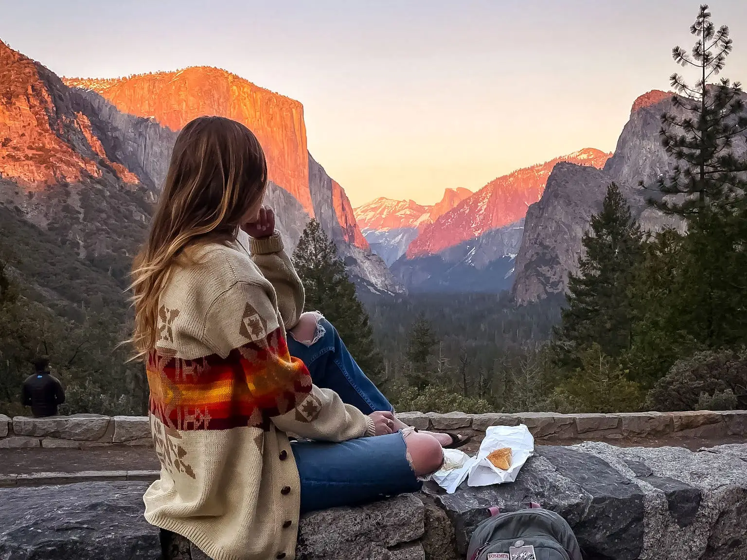 Emily sits on a wall at sunset, looking out at rock formations at Yosemite National Park. There is a bagel resting on a white paper bag next to her.