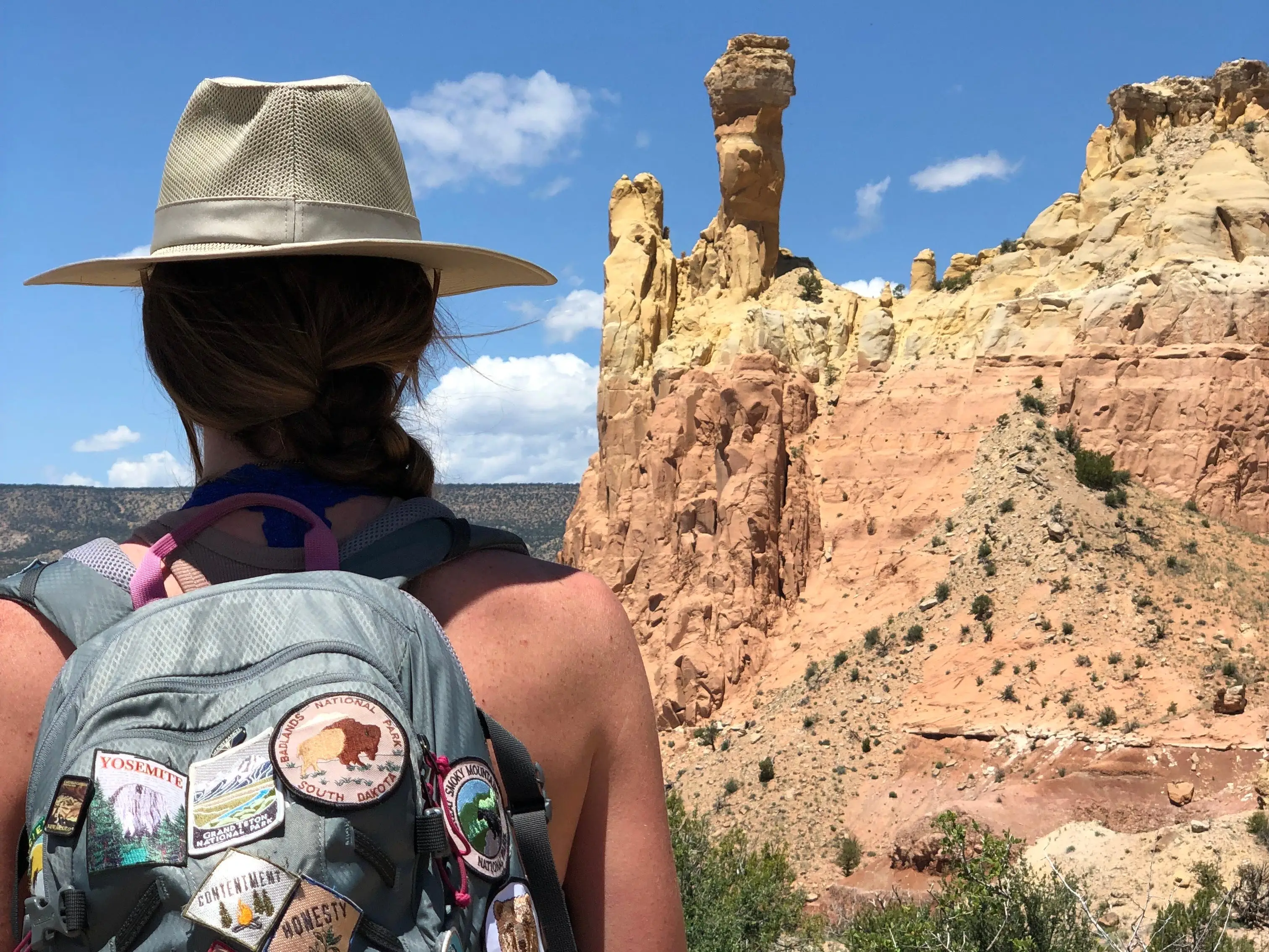 Emily, wearing a hat and a backpack covered with patches, looks out at rock formations on a sunny day.
