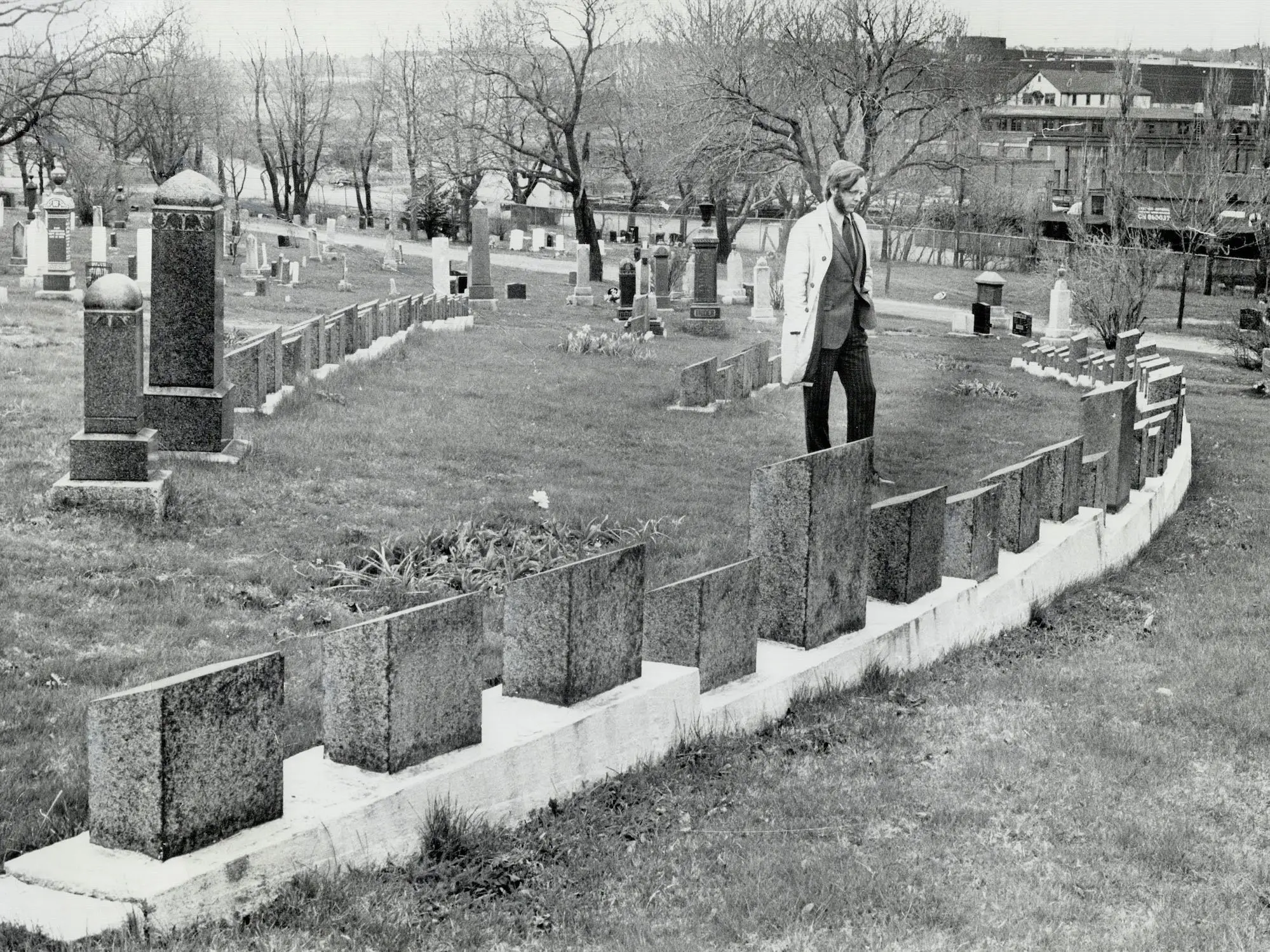 A man in a white jacket with glasses stands in front of a row of graves