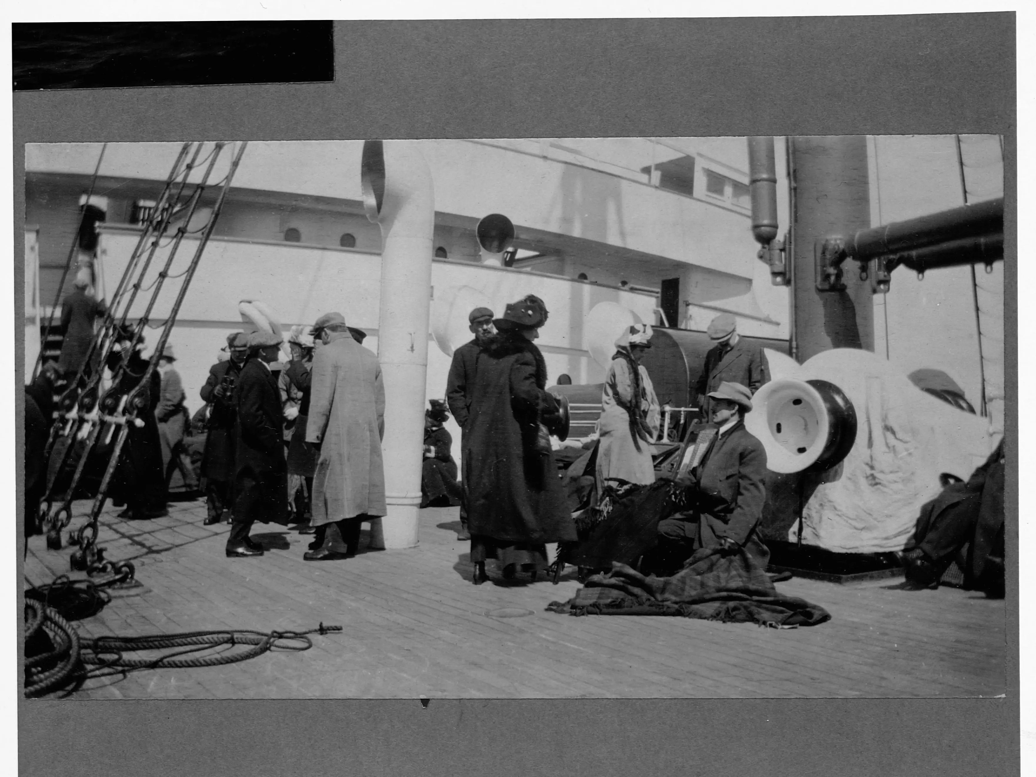 People in 1910s clothing gather on a ship's deck with ropes and equipment nearby