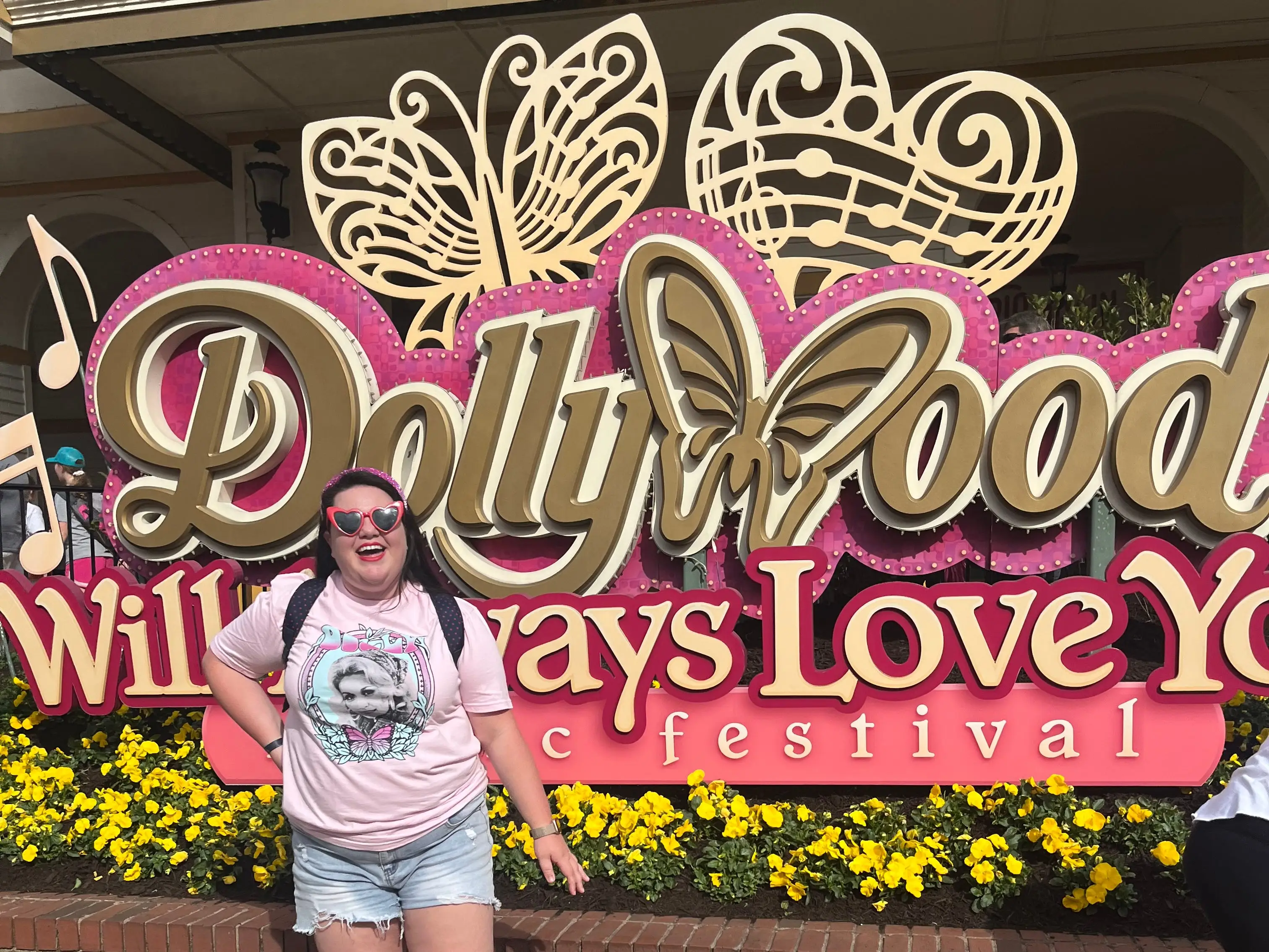 A woman wearing a Dolly Parton T-short and heart-shaped sunglasses standing in front of a Dollywood sign.