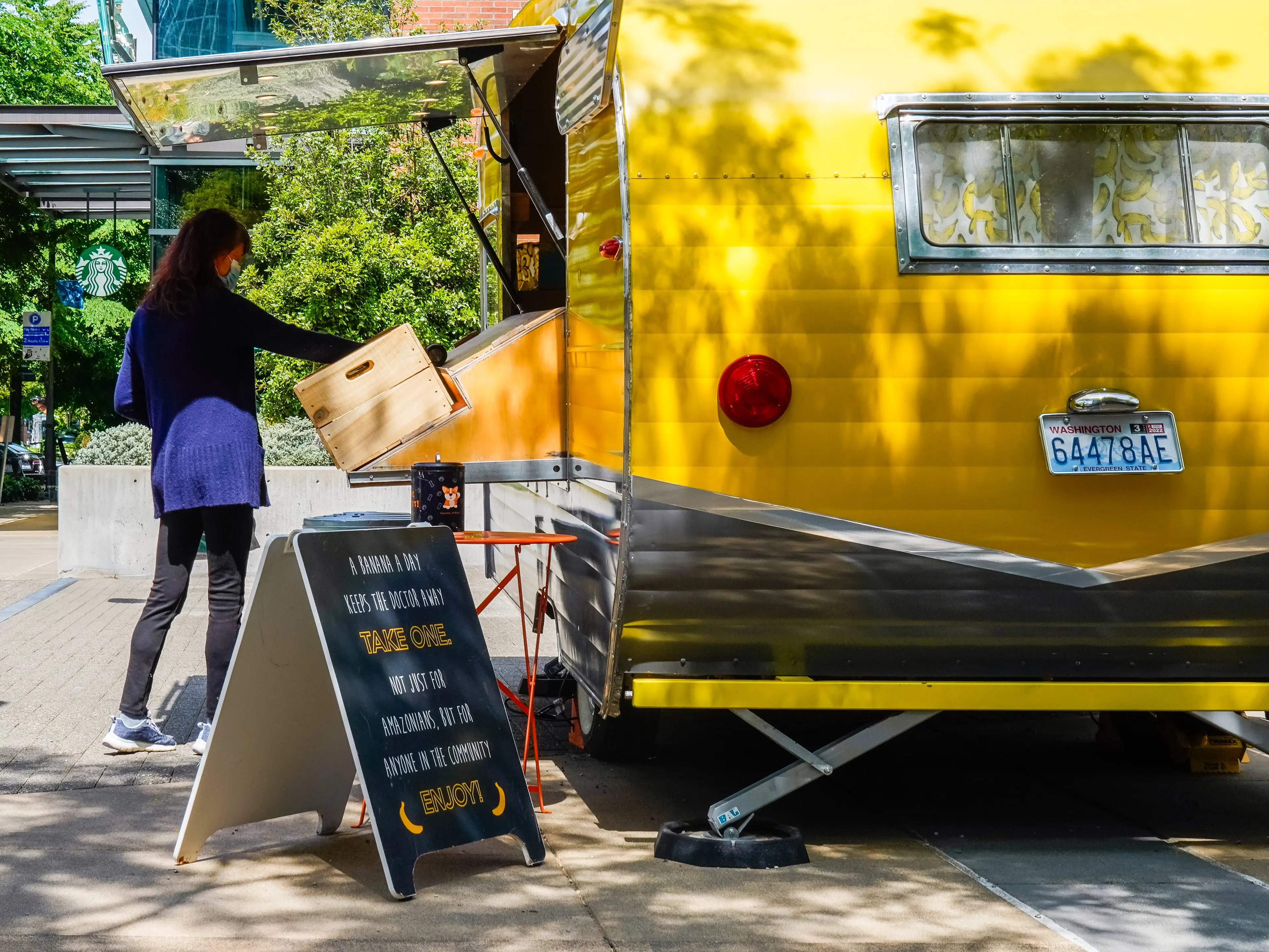 A woman takes a banana from a yellow trailer with trees and buildings in the background