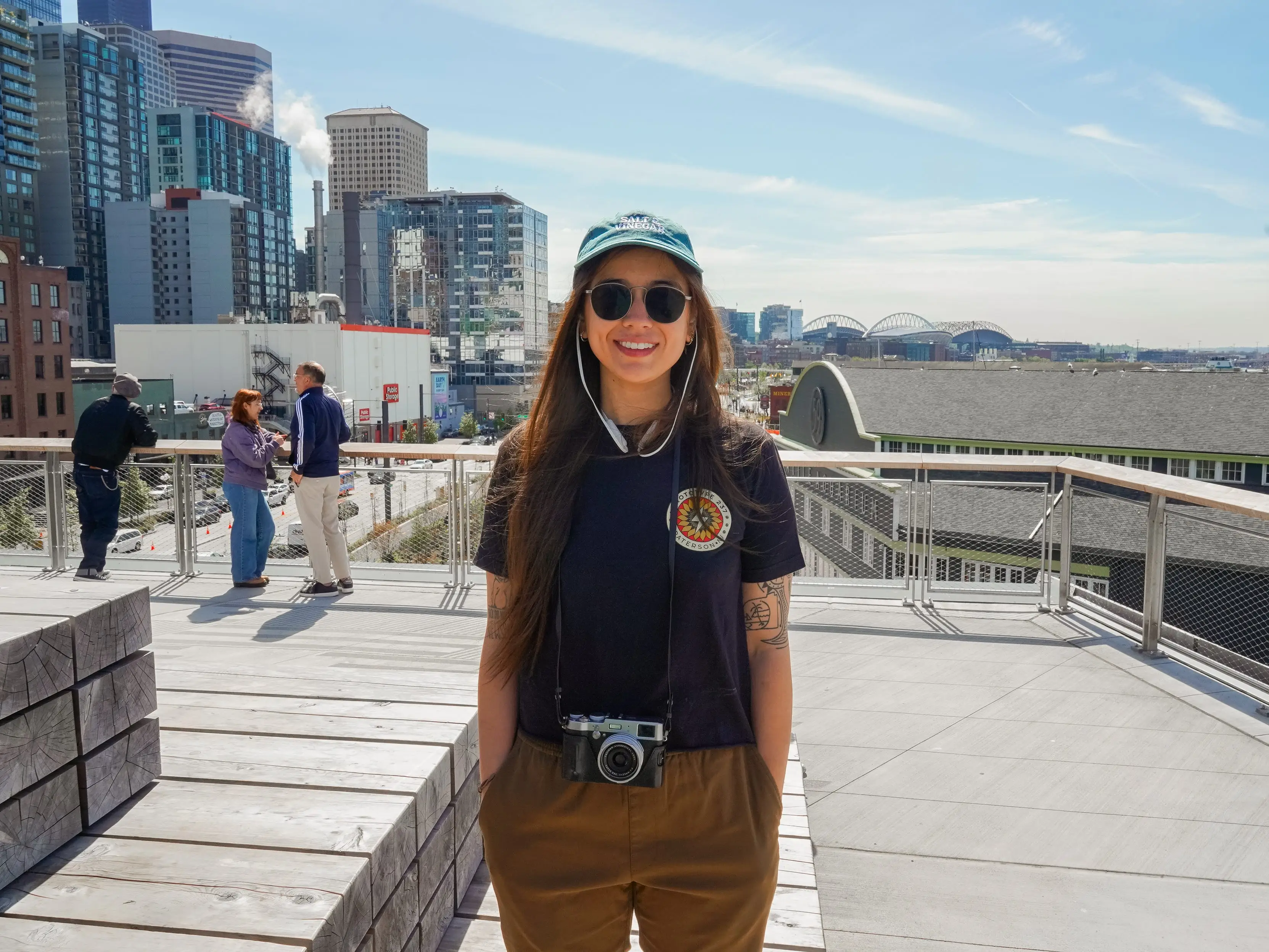 The author, wearing a camera around her neck, stands smiling on a Seattle rooftop with a view of the city behind her.