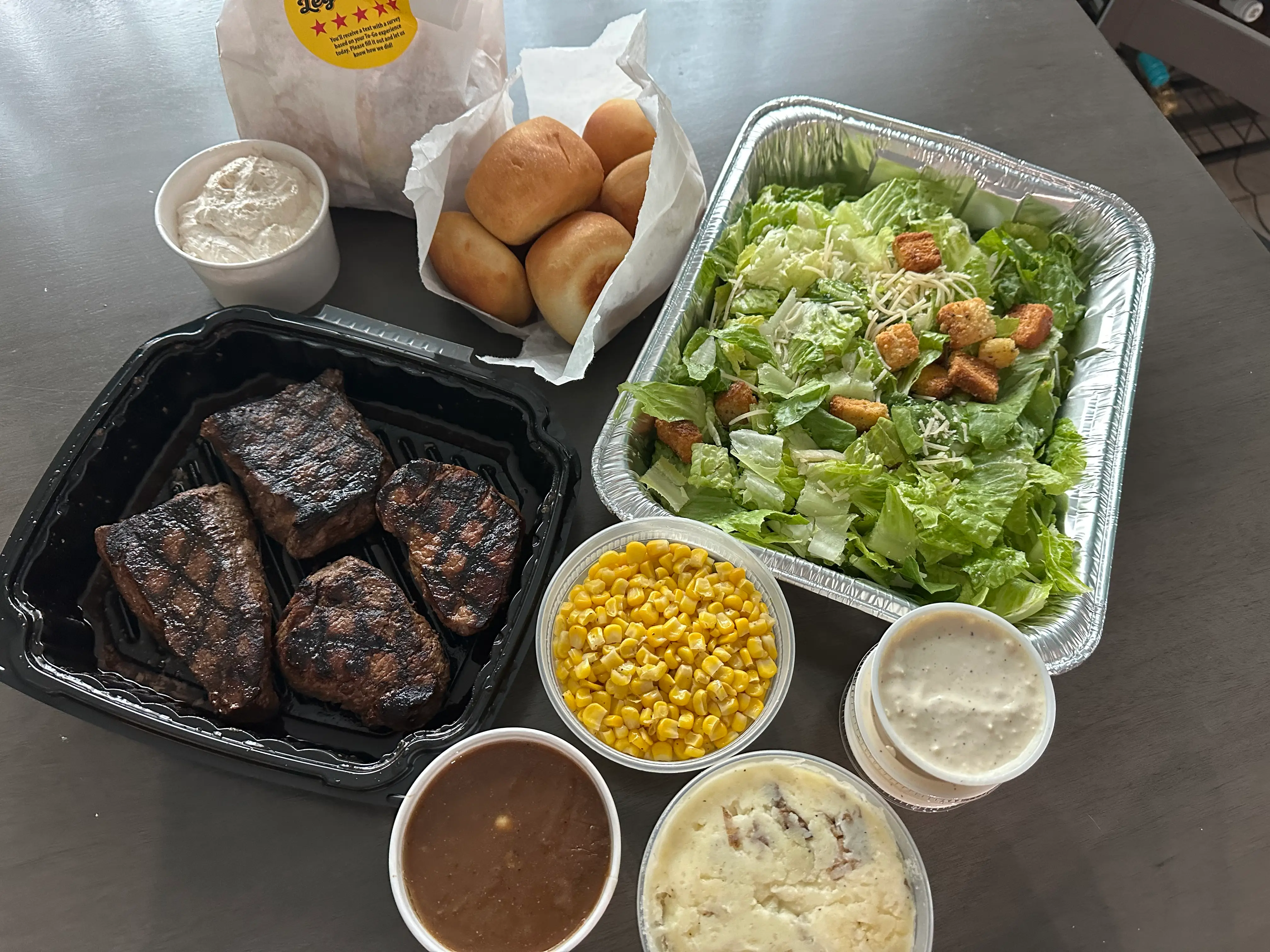 Four steaks, large salad, bag of rolls, and other sides from Texas Roadhouse on table