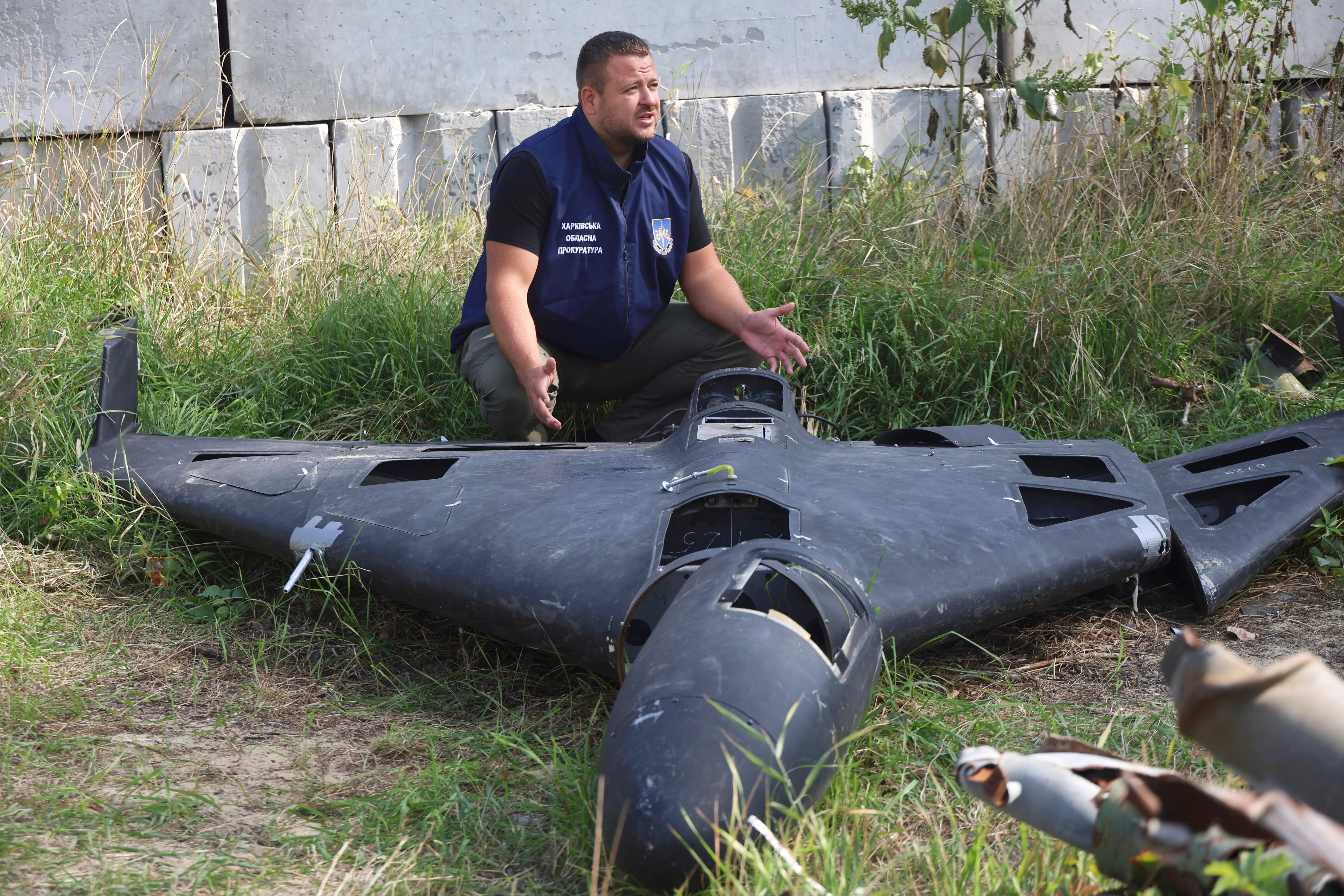 A man in a blue vest squats beside a large black-colored drone that is damaged and lying on a grassy ground