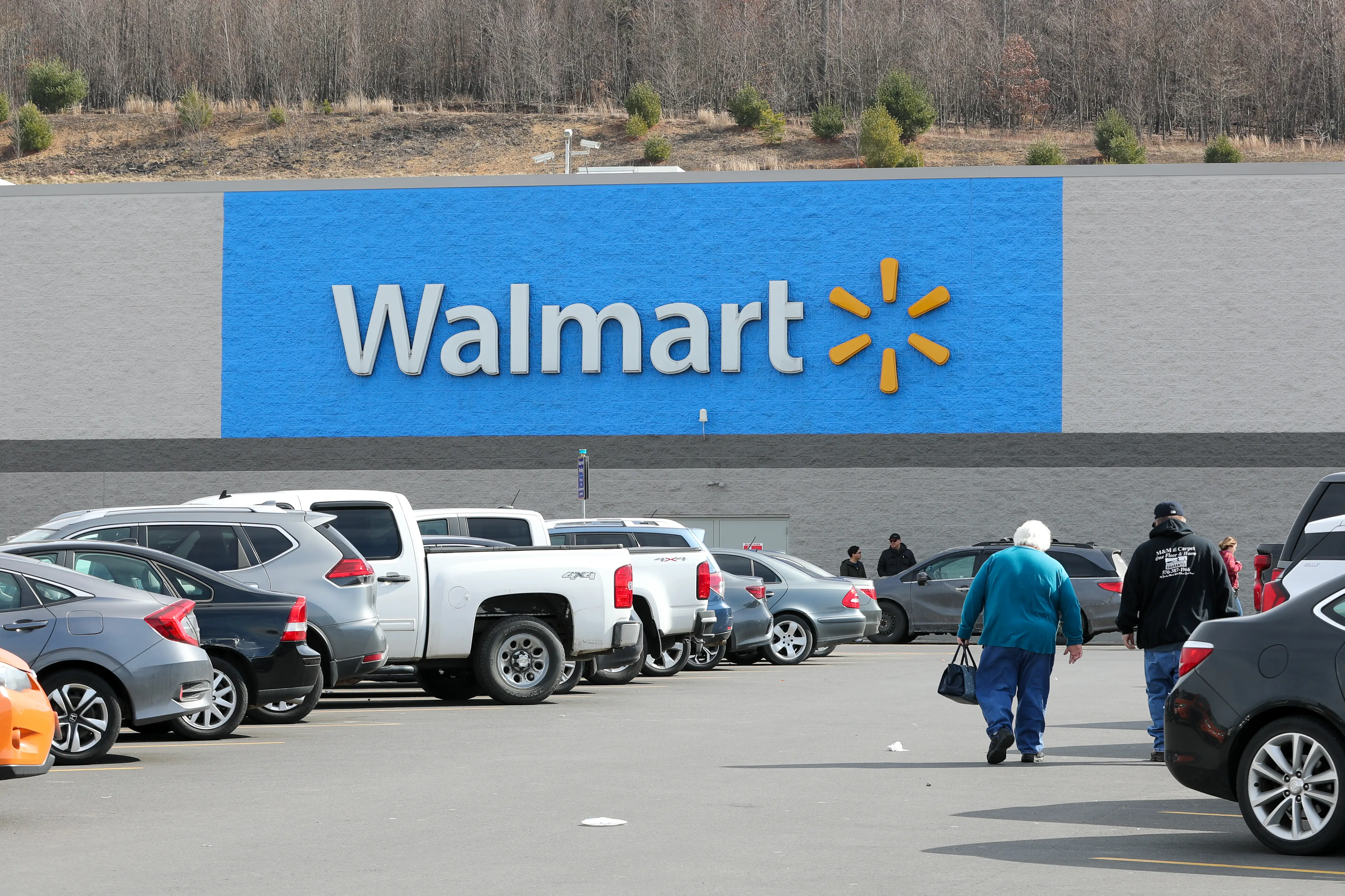 Customers walk through a Walmart parking lot, with cars in the foreground and a white-and-yellow Walmart sign mounted on a wall of the store painted blue