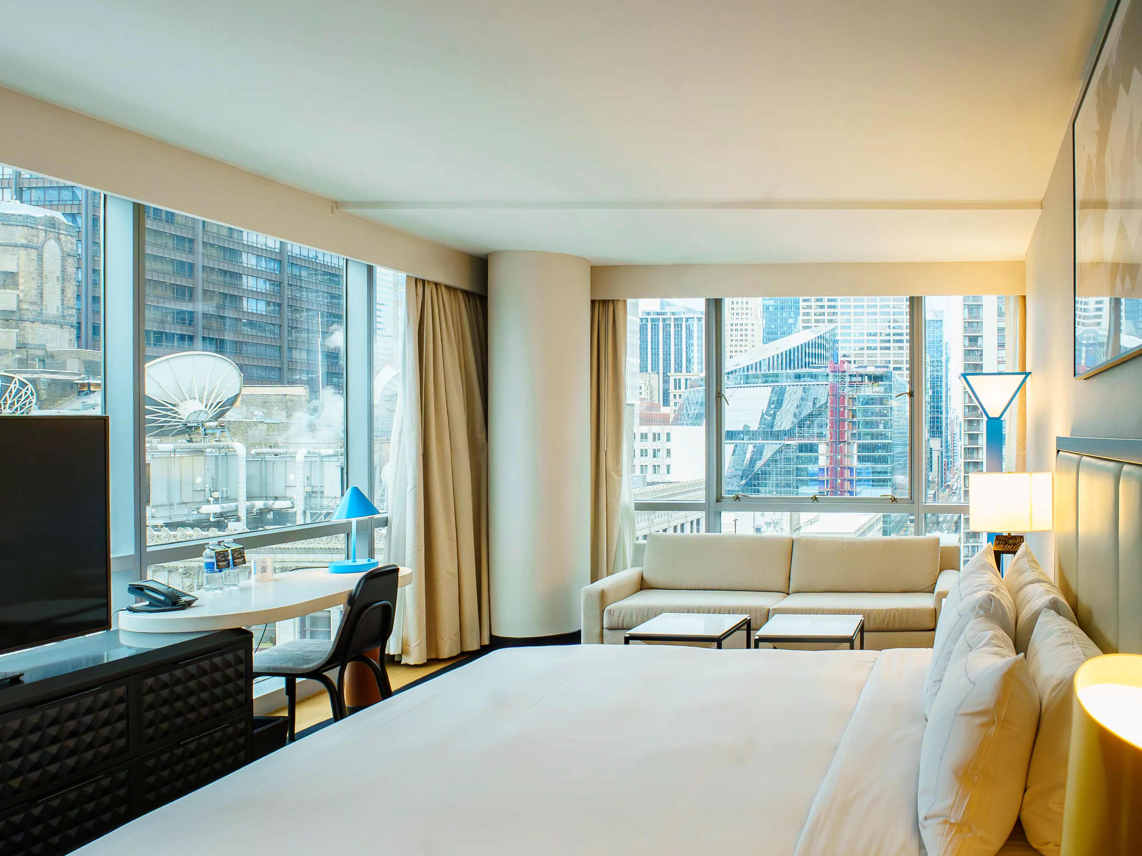 Inside a room at the Wit, theres a bed on the right, a TV and desk on the left, a couch in the back, and wide windows on. both walls