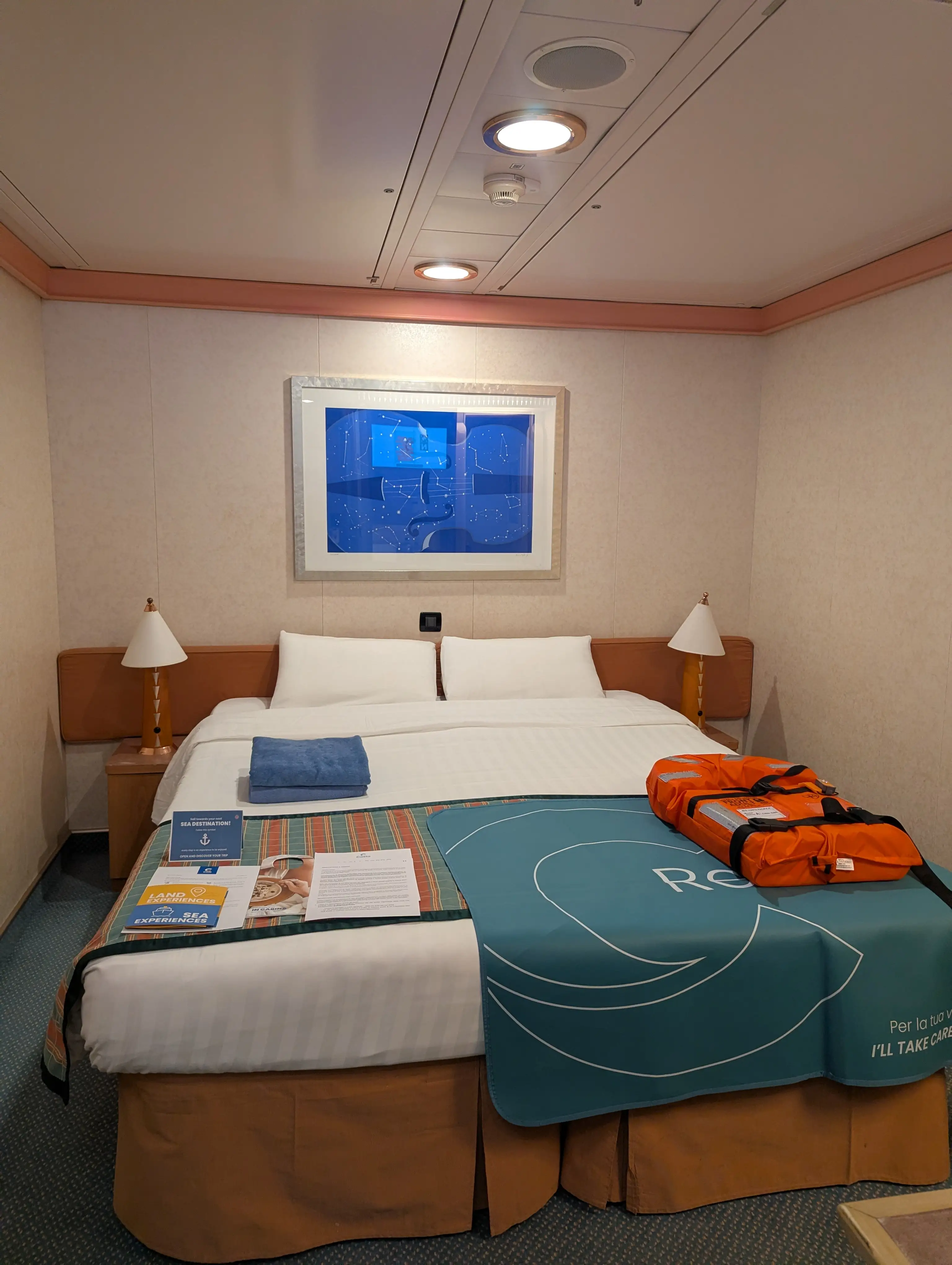 Interior cabin on Costa cruise with bed, two lamps on either side, simple wallpaper