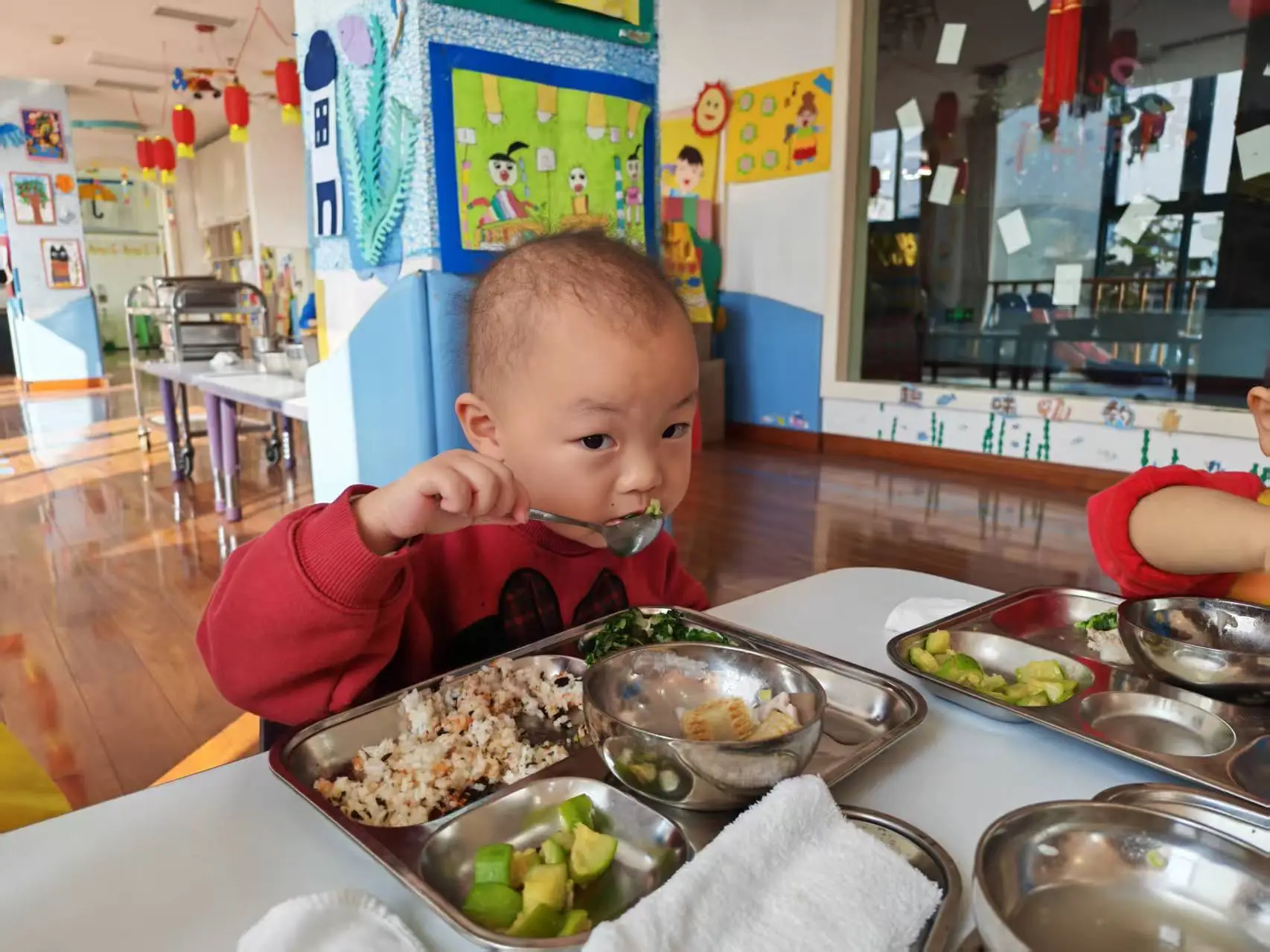A toddler eating school lunch fom silver bowls on a tray.