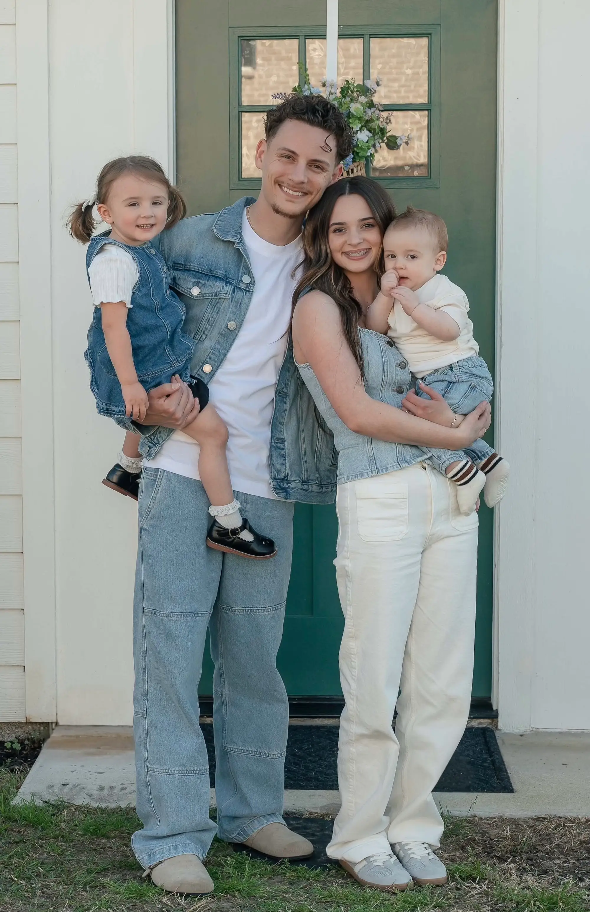 A couple stands in front of a green doorway holding their two children.