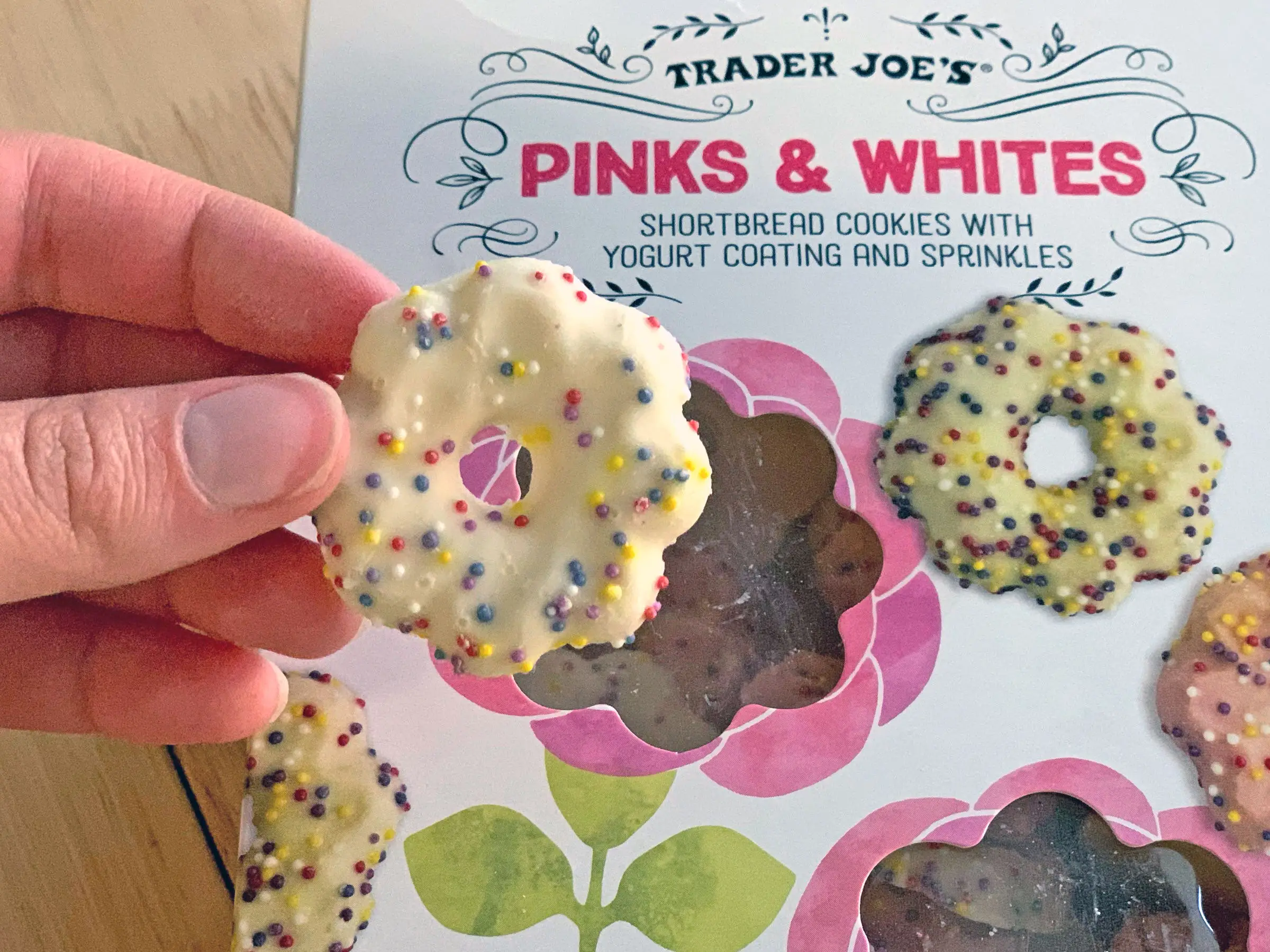 A hand holding an iced shortbread cookie covered in icing and sprinkles. Behind it is the box the product came in.