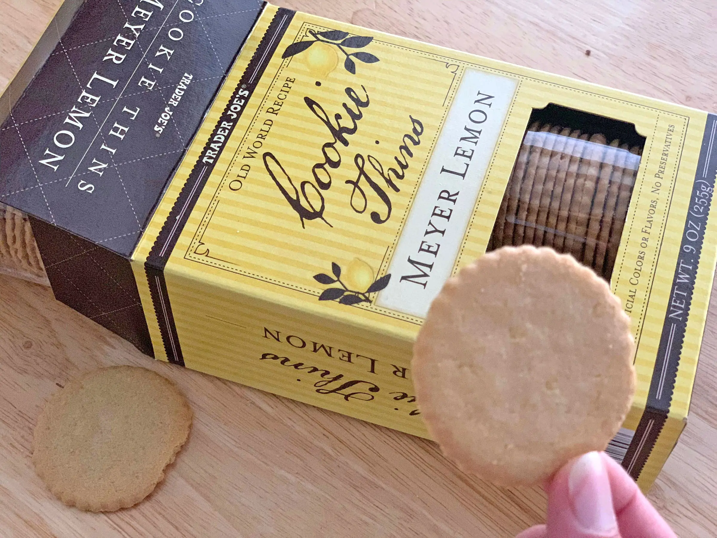 A hand holding a Trader Joe's Meyer lemon cookie thin over the box the product came in.