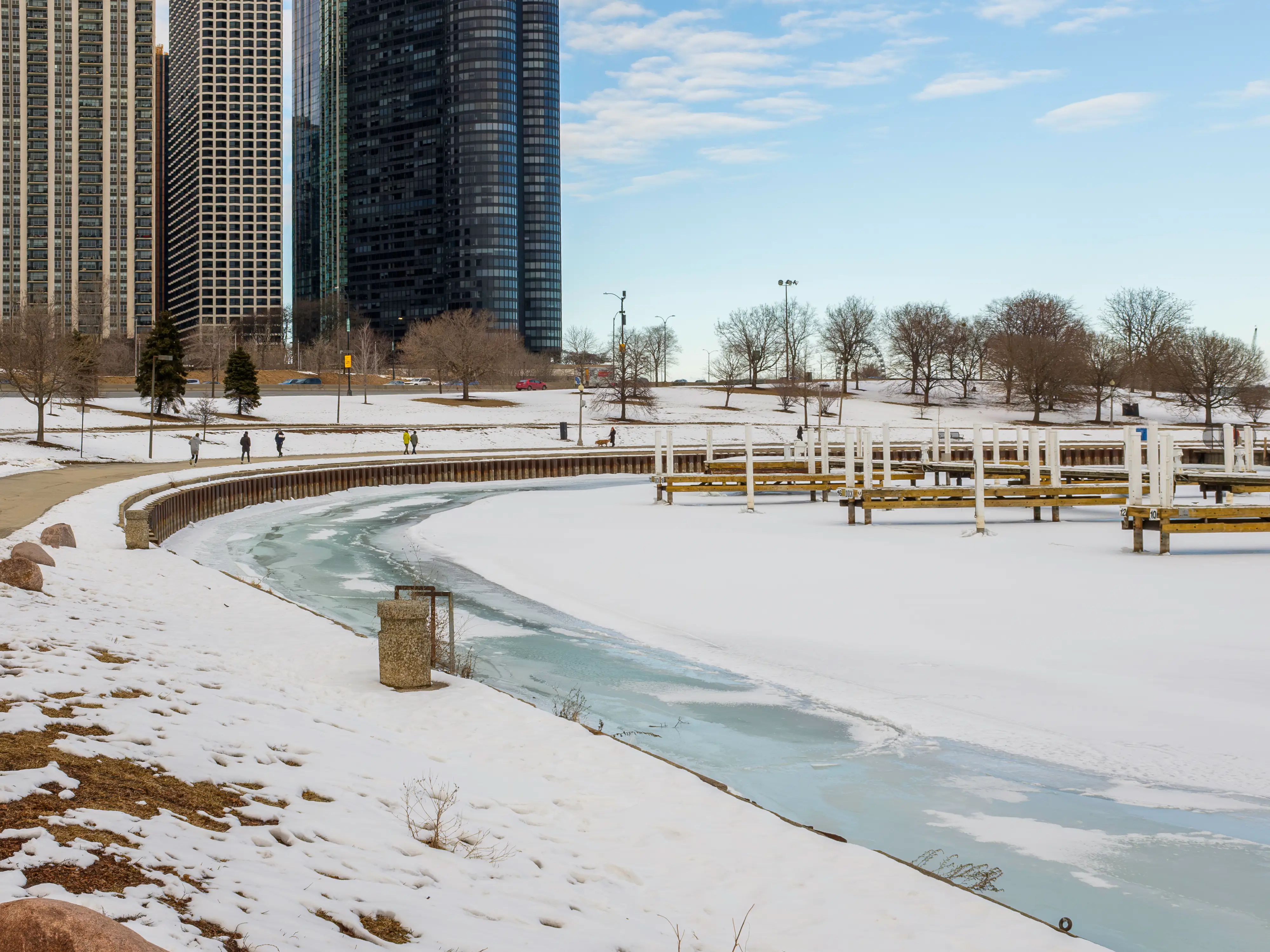 A curved edge of a frozen lake with a pathway and buildings on the left