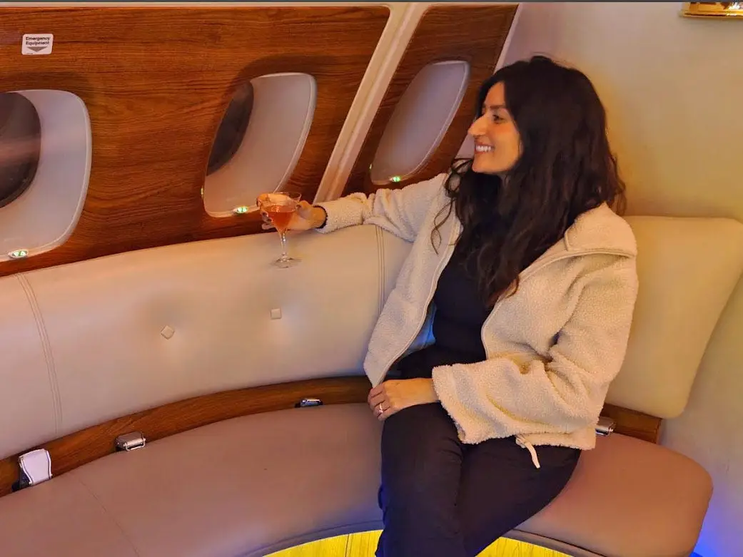 Alyssa sits on a plush bench with a cocktail and looks out the window of a plane.