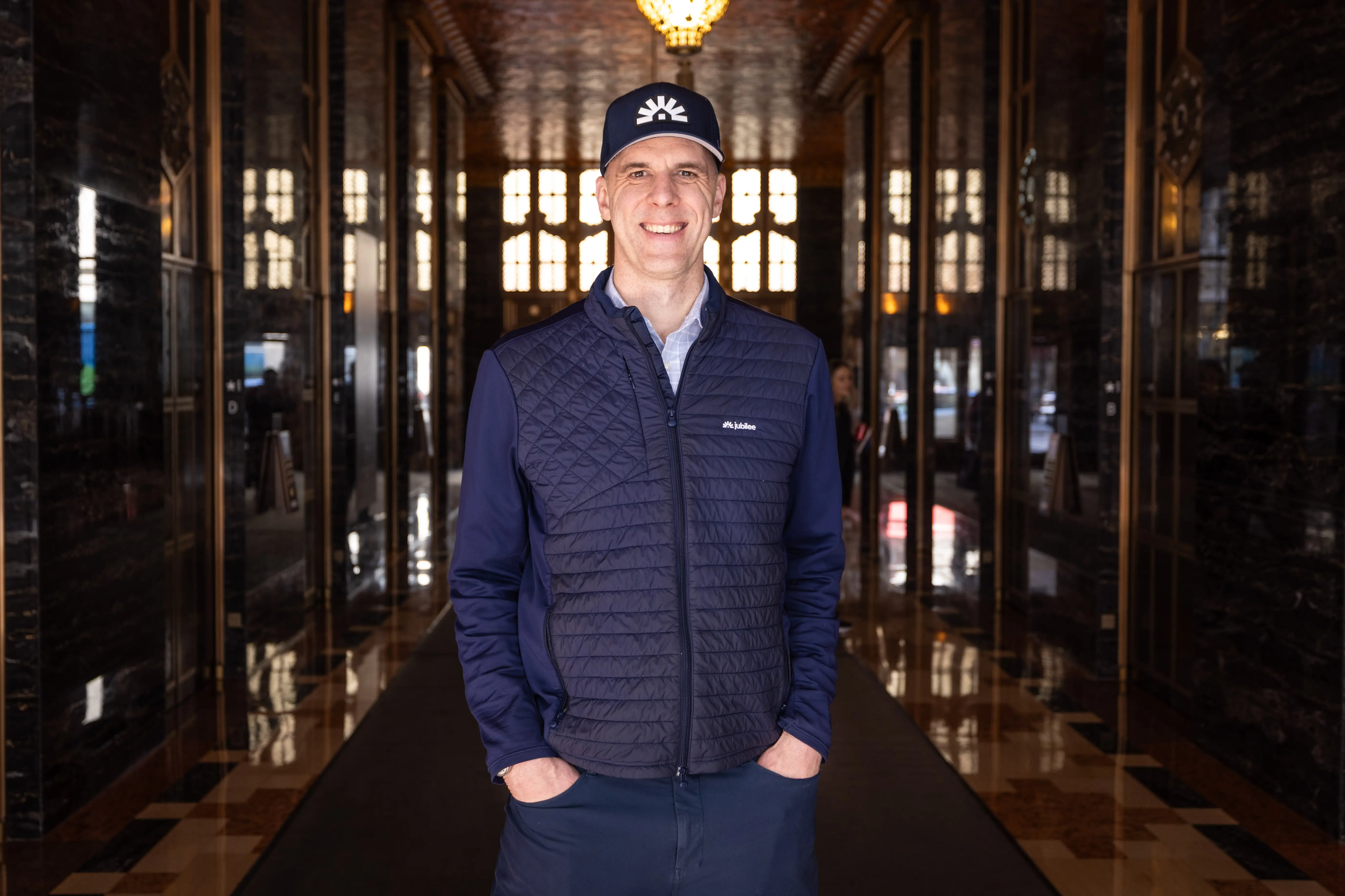 Brian Elbogen, the founder and CEO of Jubilee Homes, stands in a hallway wearing a hat and vest with the company logo