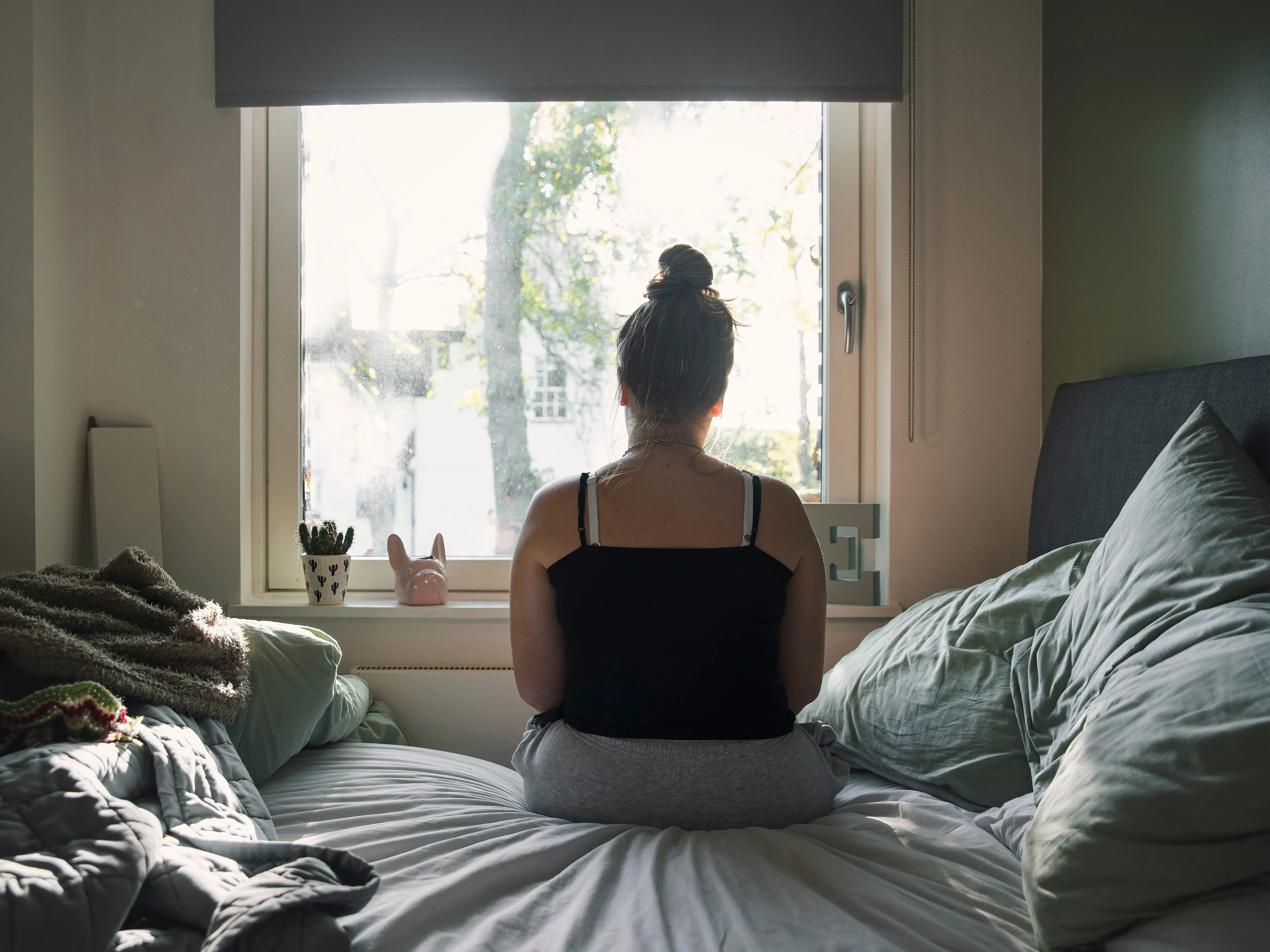 A woman sits on a bed and looks out a window