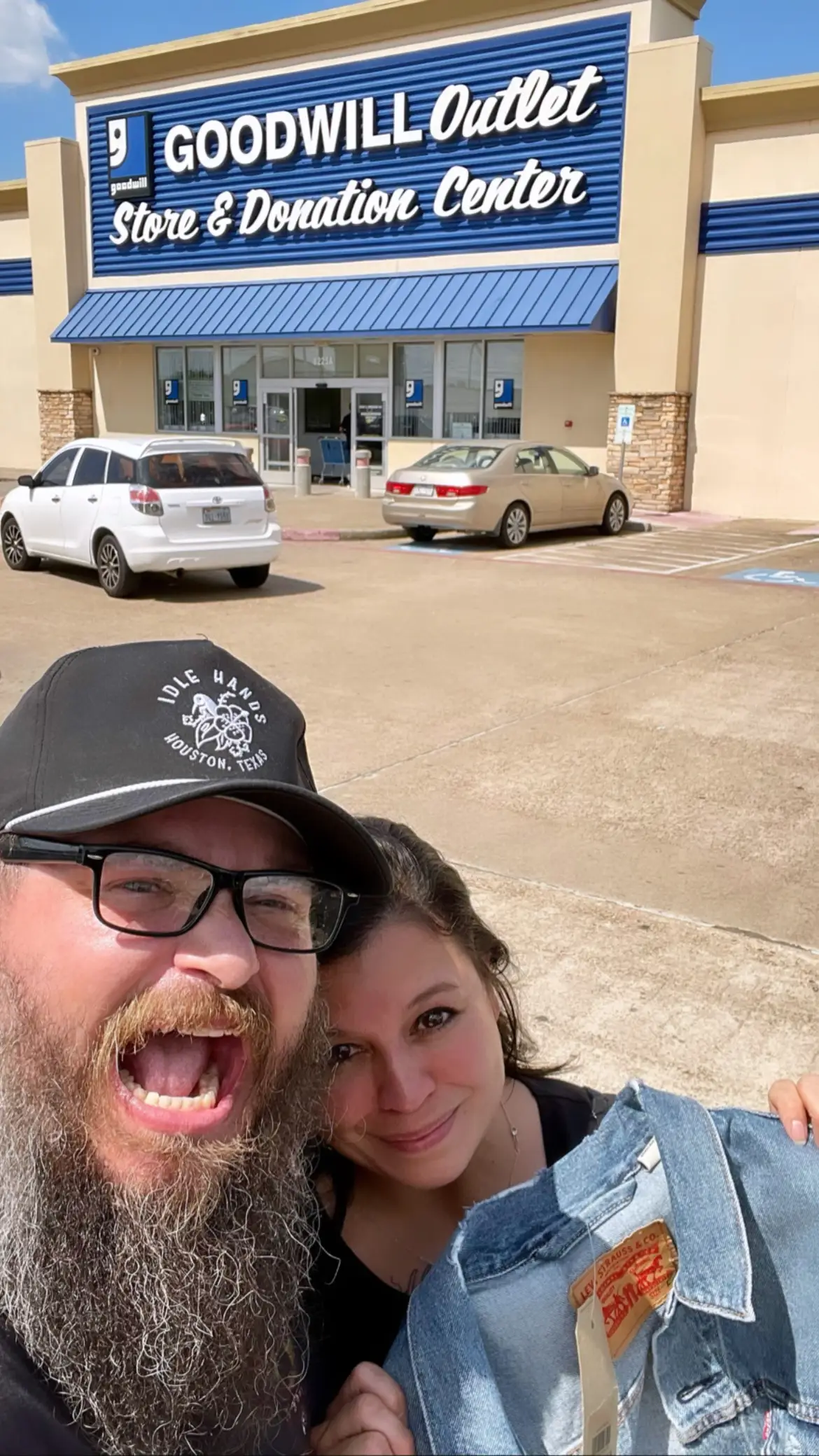 A couple hold up one of their finds in he parking lot of a Goodwill. She's smiling, he's making a funny face.