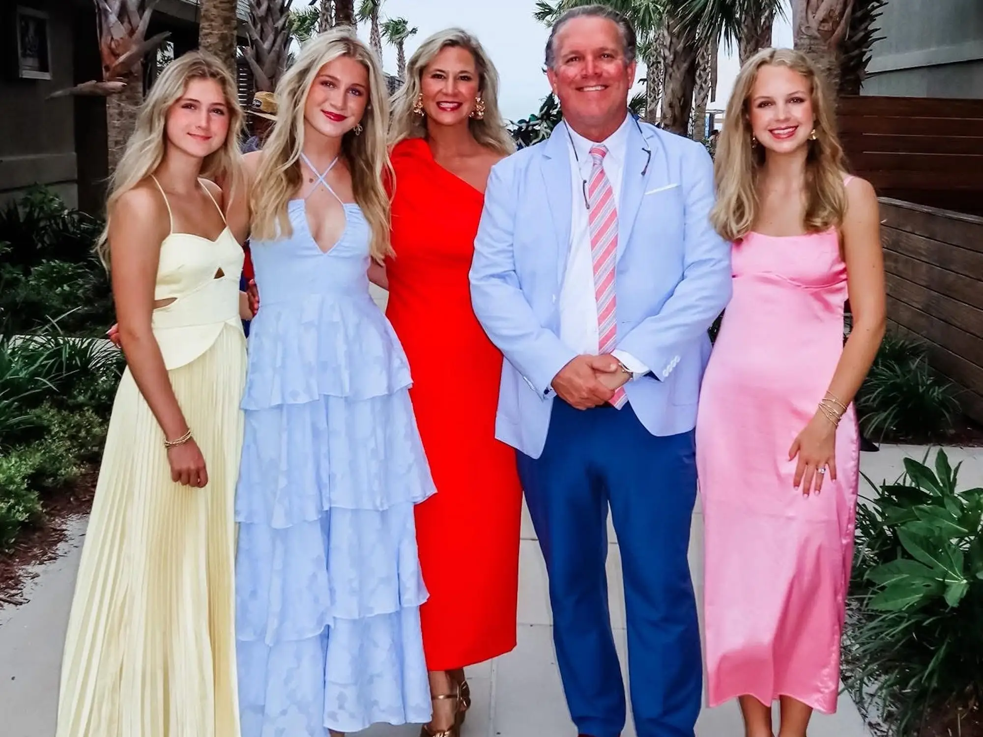 A family of five stands together between palm trees.