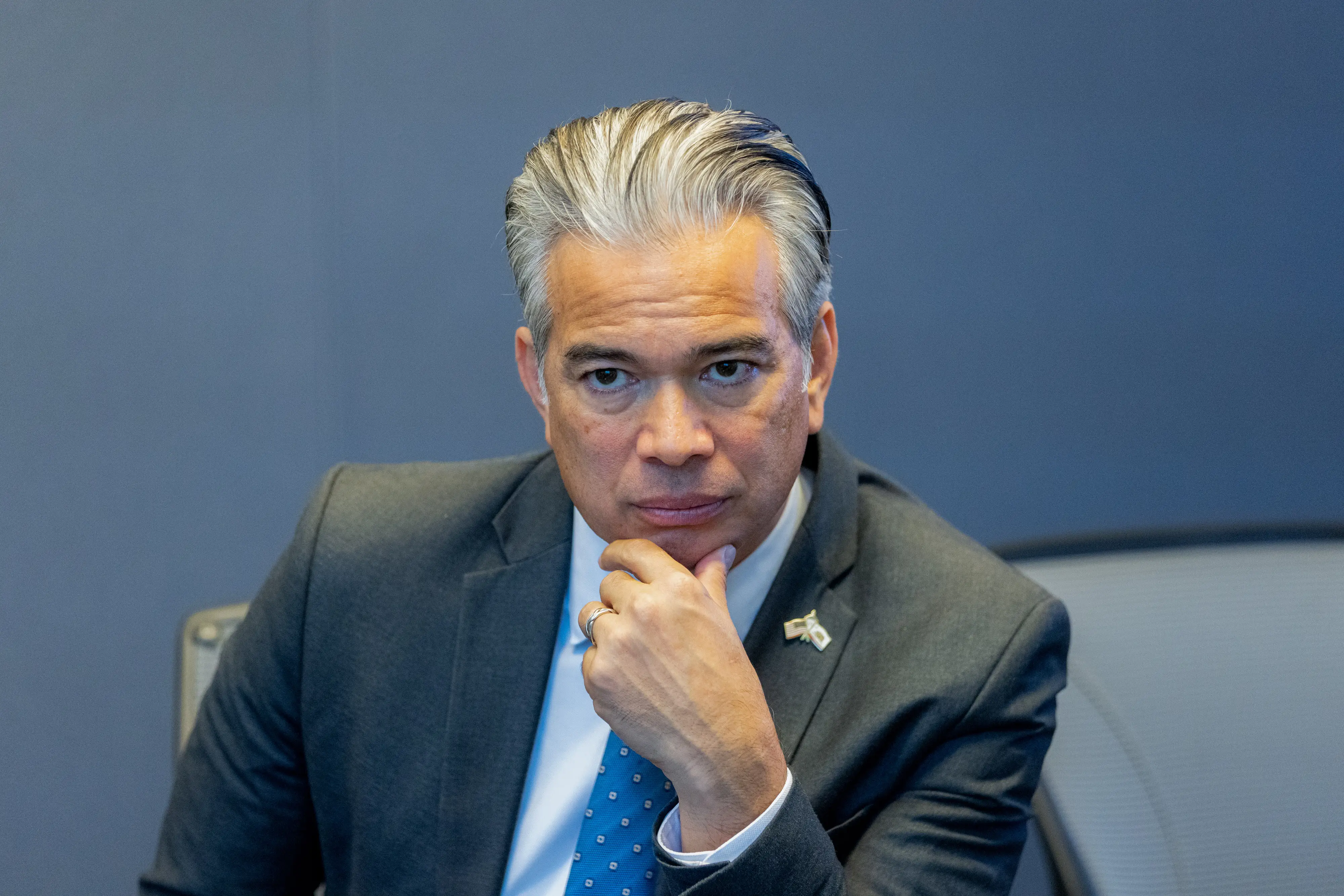 Rob Bonta, California's attorney general, wearing a suit and blue tie, holds his hand to his chin while seated at a table looking toward the camera
