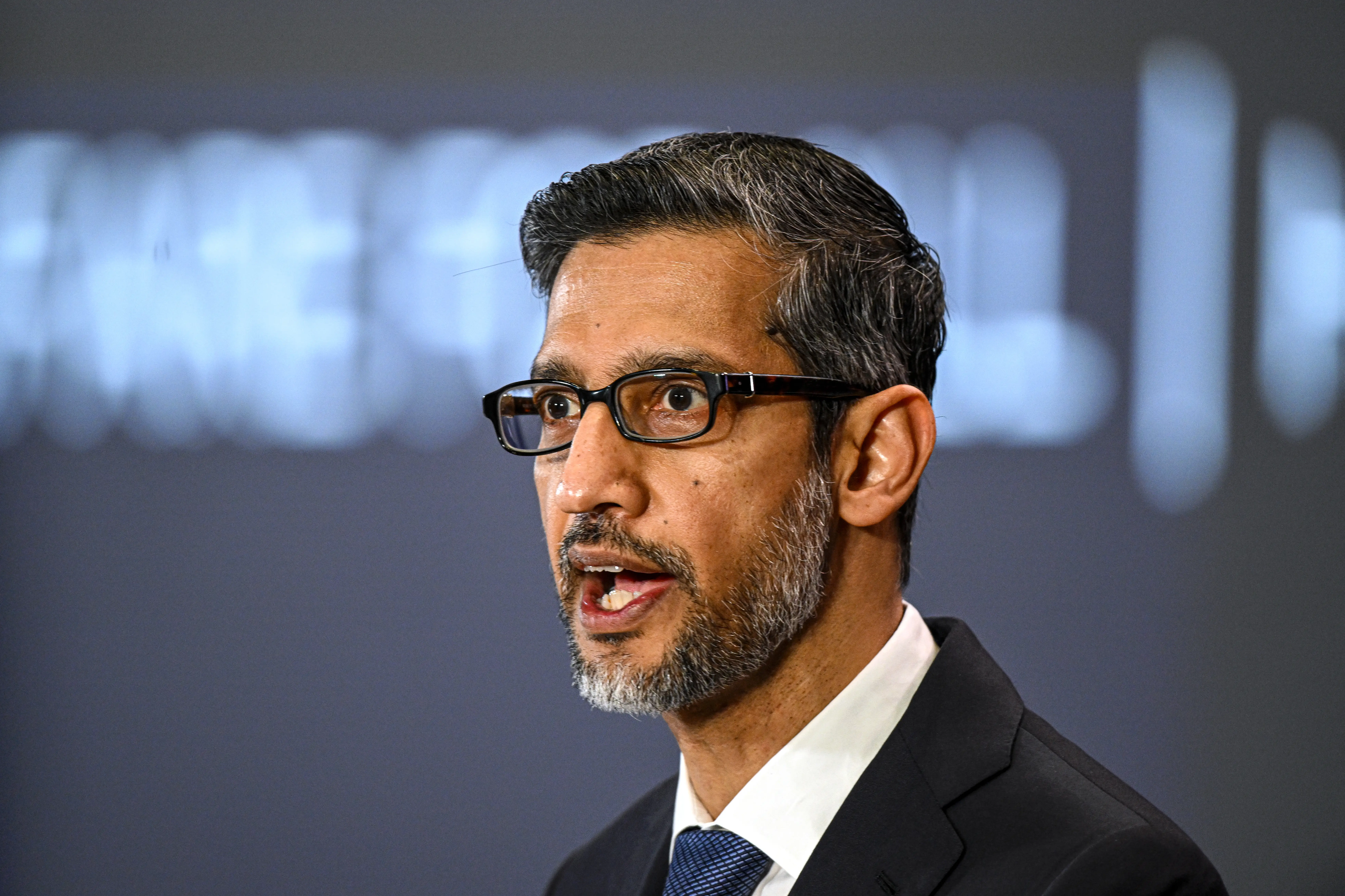 Sundar Pichai wears a black suit and navy tie on a dark background.