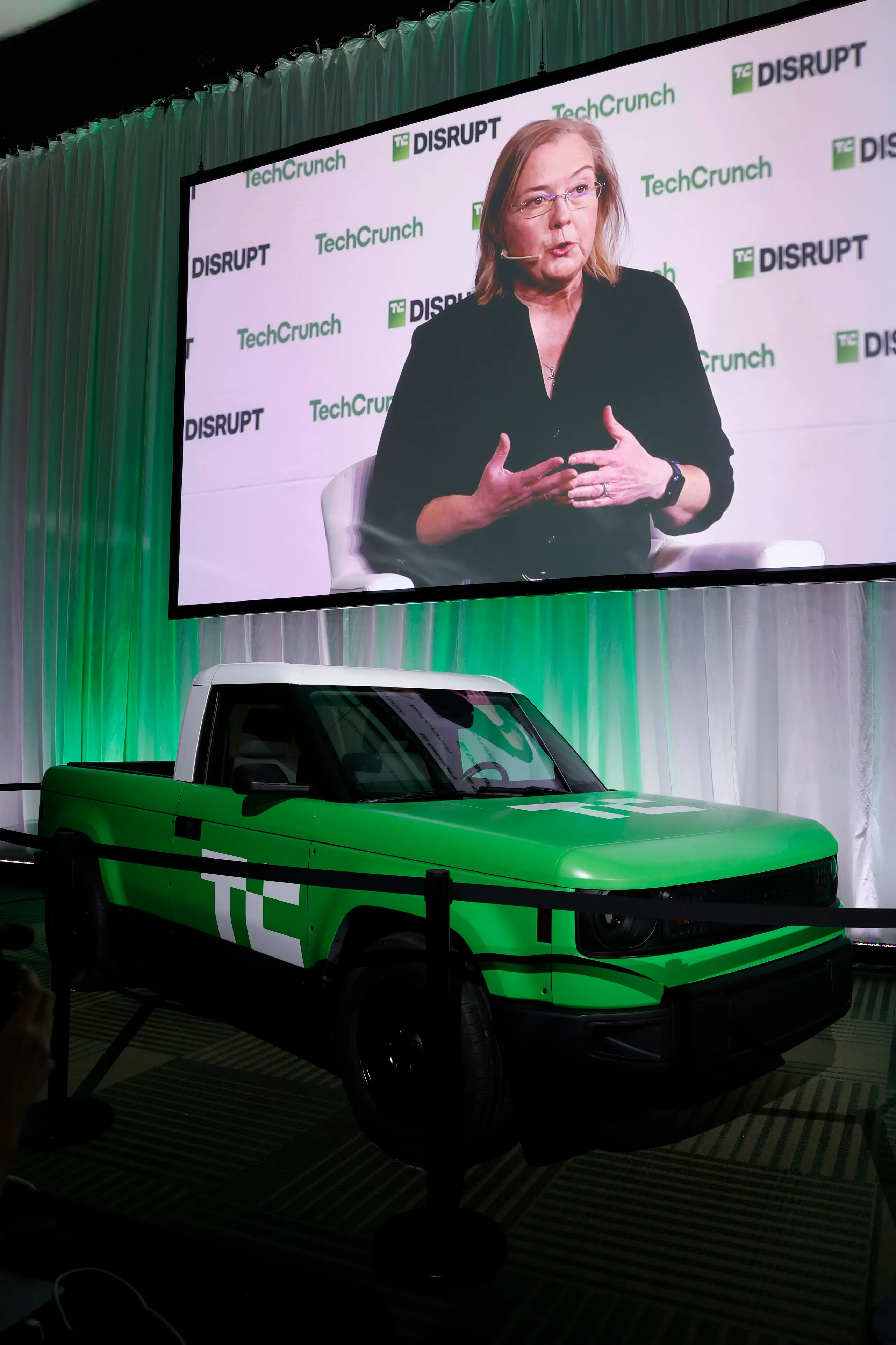 A Slate truck, wrapped in green with a white top, is parked behind a stanchion. Chris Barman, the company's former CEO, is projected on a screen behind the truck.