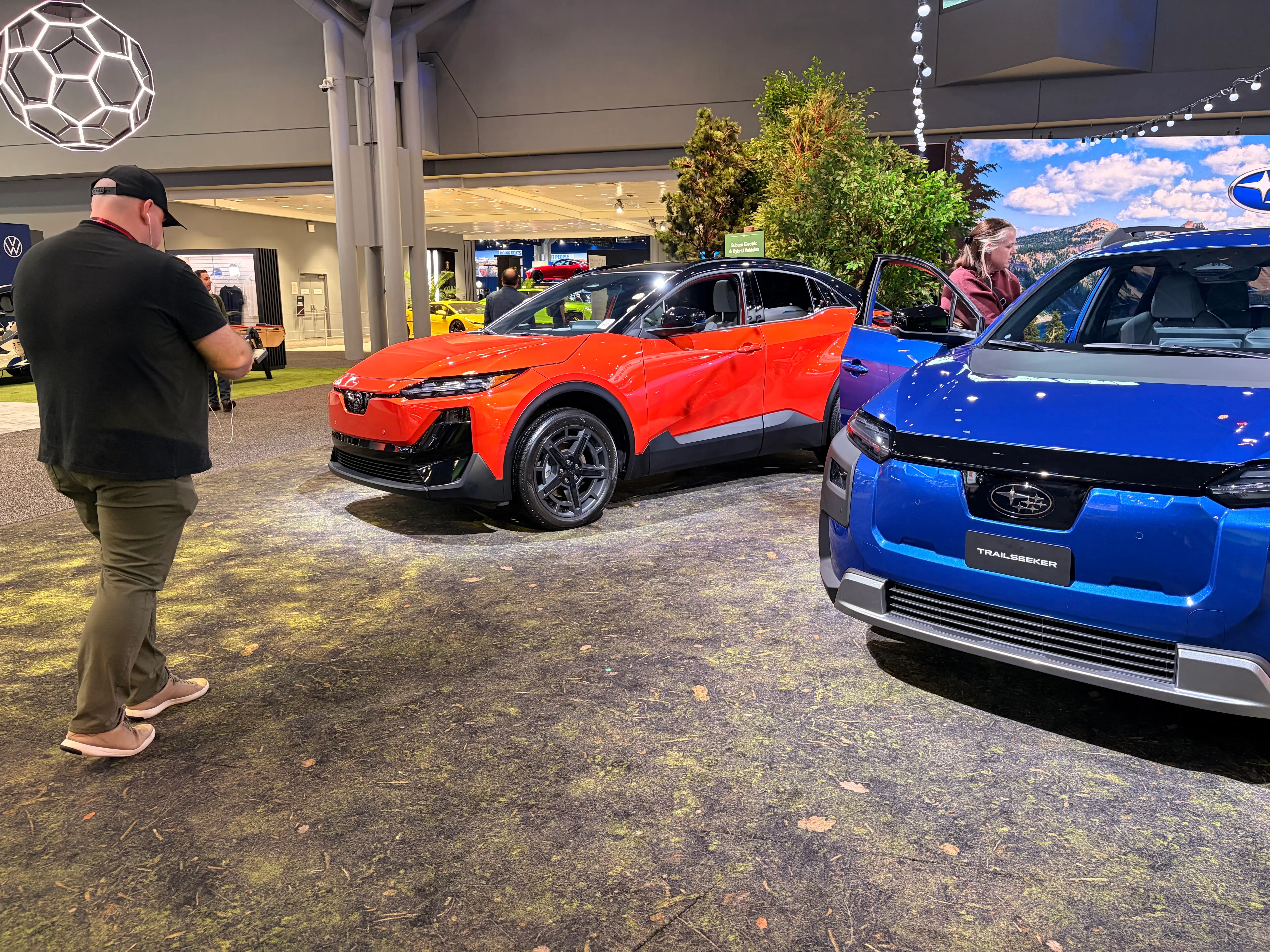 An orange Subaru Uncharted is parked next to a blue Subaru Trailseeker. Photographers are snapping pictures of both cars.