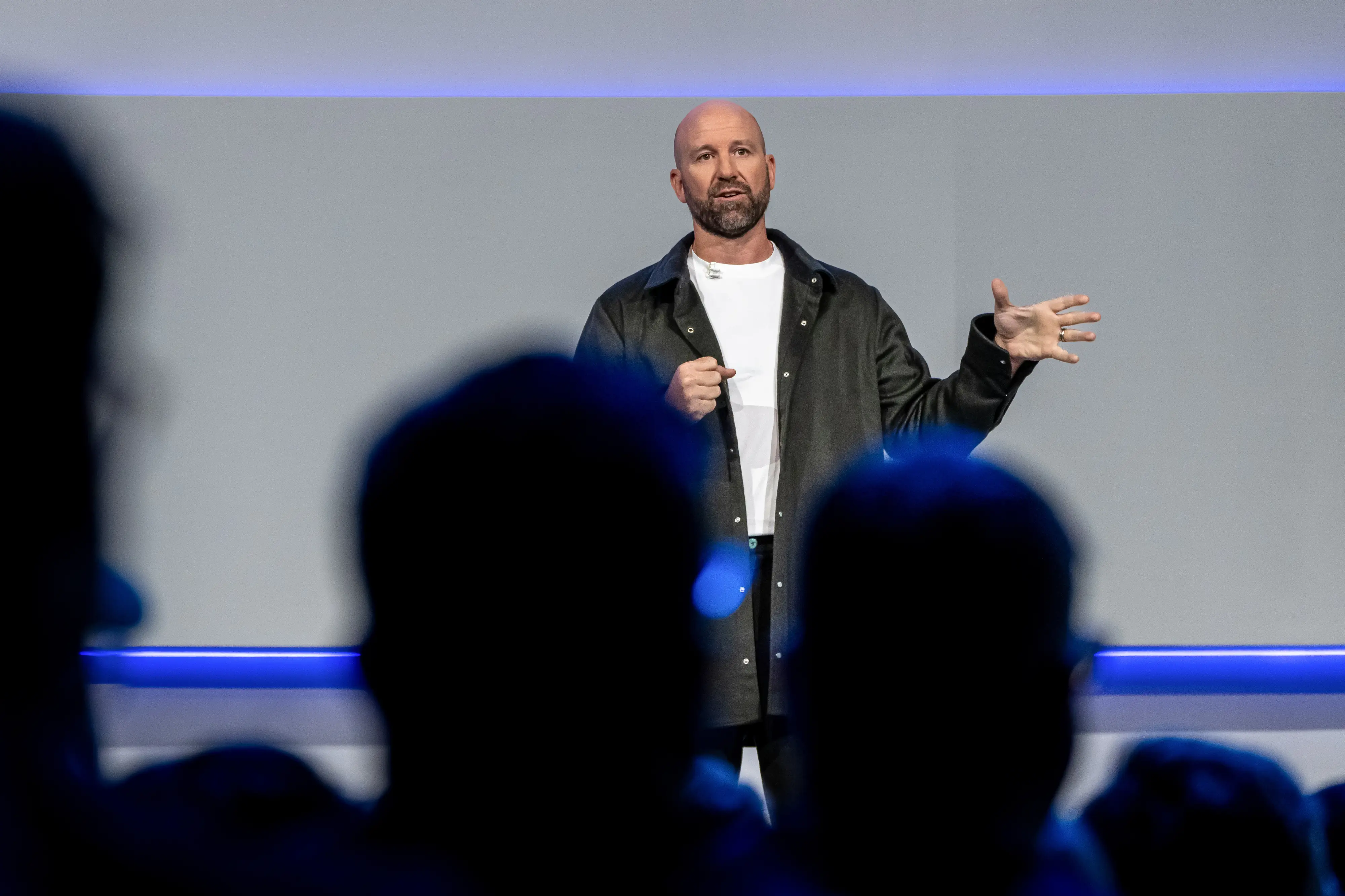 Andrew Bosworth, Meta's CTO, stands on stage in a long gray jacket and a white shirt. His left hand is splayed.