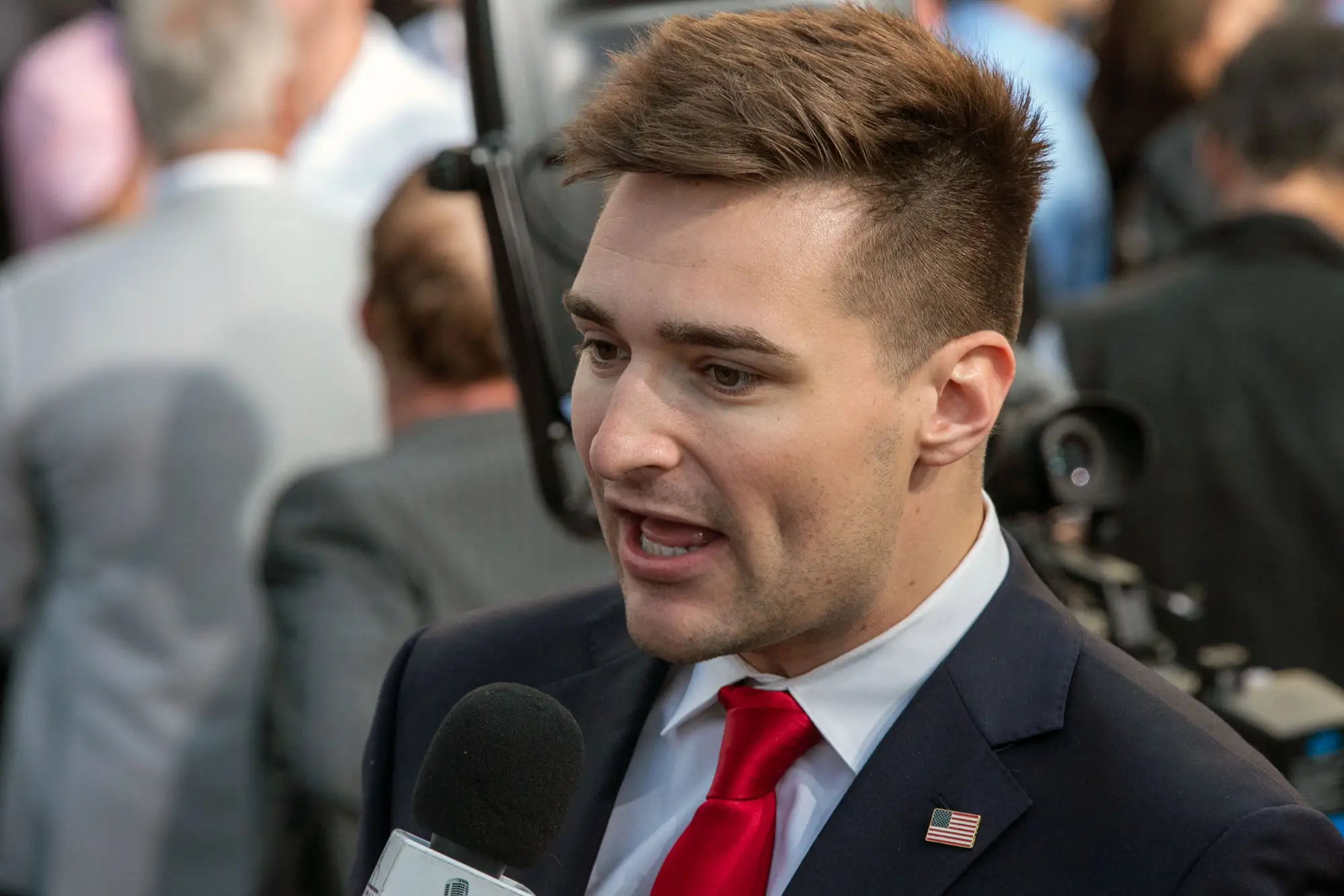 Alex Bruesewitz speaks into a microphone, wearing a suit with an American flag pin on its lapel.