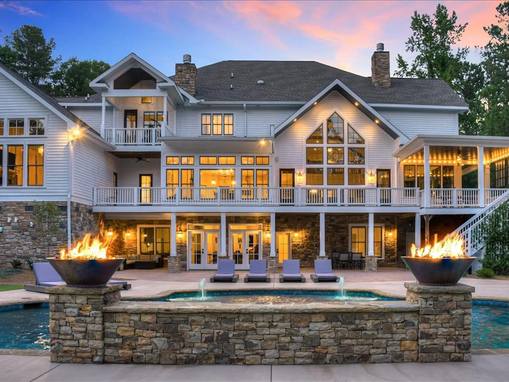 The back of a house with a pool in front of it at sunset. Firepits frame the pool.