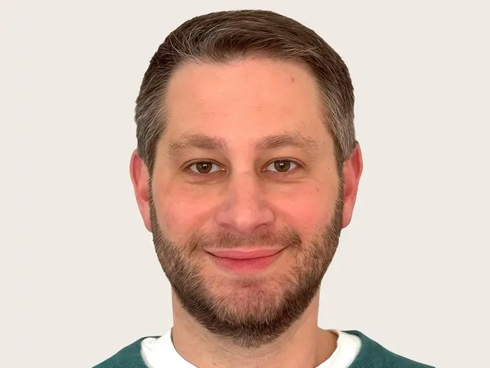 A smiling man with short brown hair and a trimmed beard faces forward against a plain background.