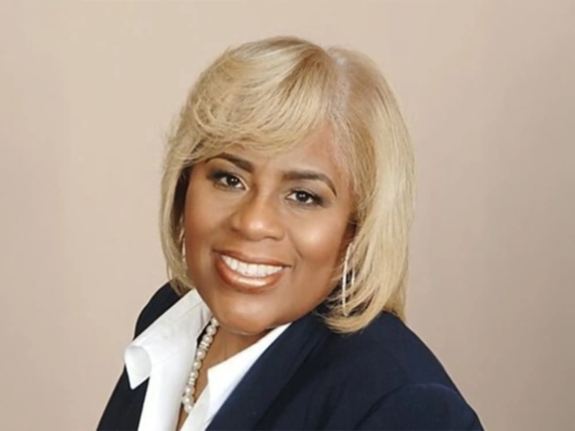 Pamela Price smiling in a headshot with a tan background, a dark navy blue blazer, a white blouse, and pearls.
