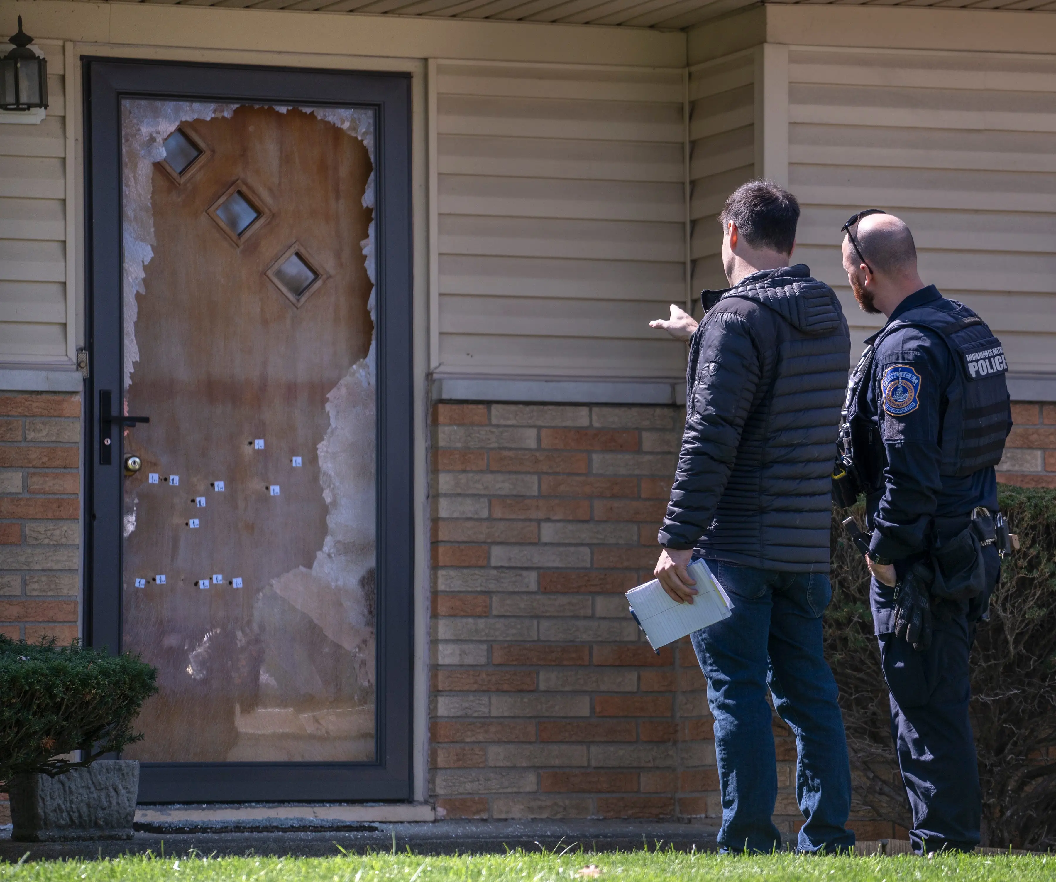 Two men in front of a door with bullet holes.