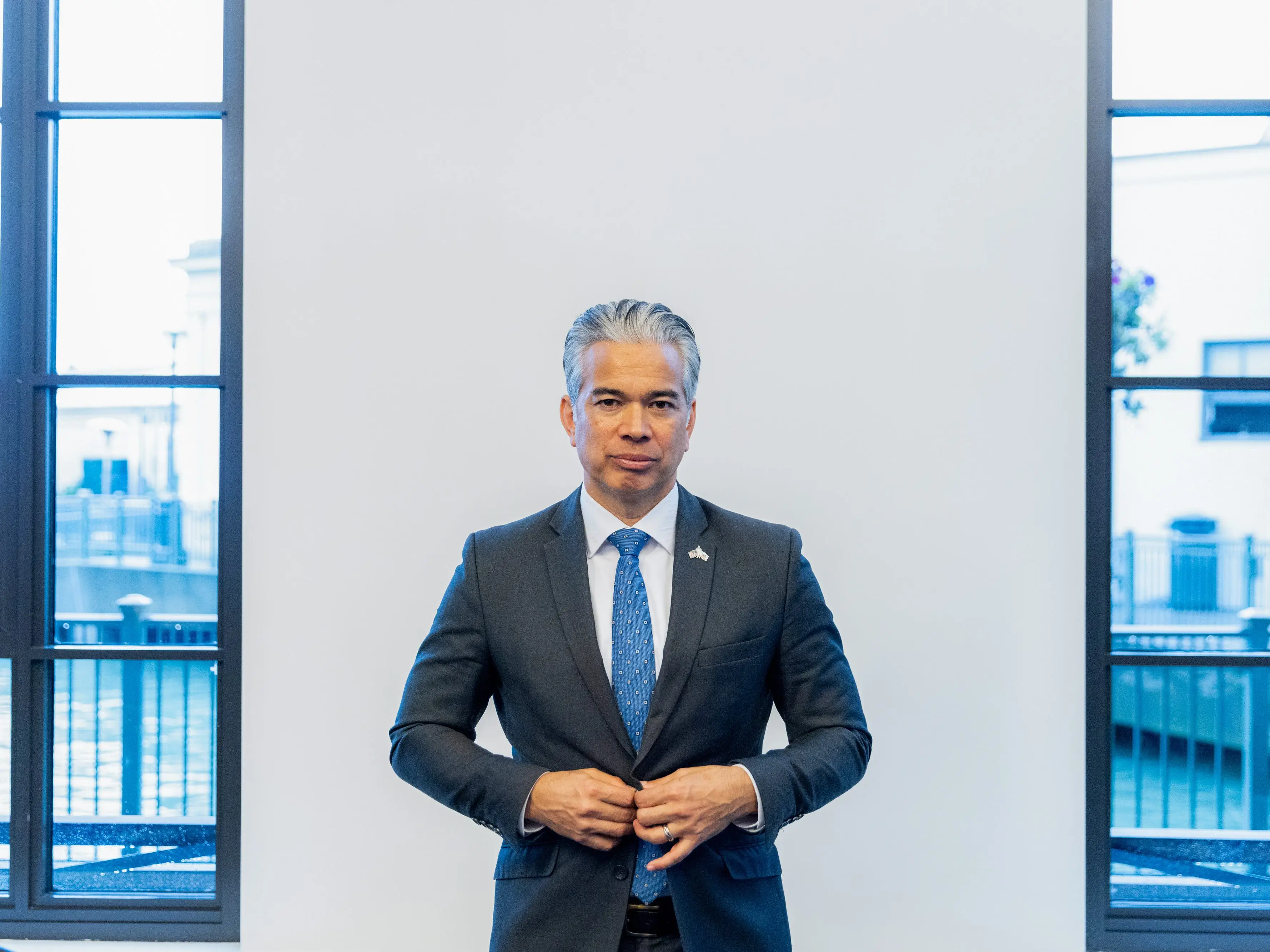 Rob Bonta buttons a suit jacket standing by a window.
