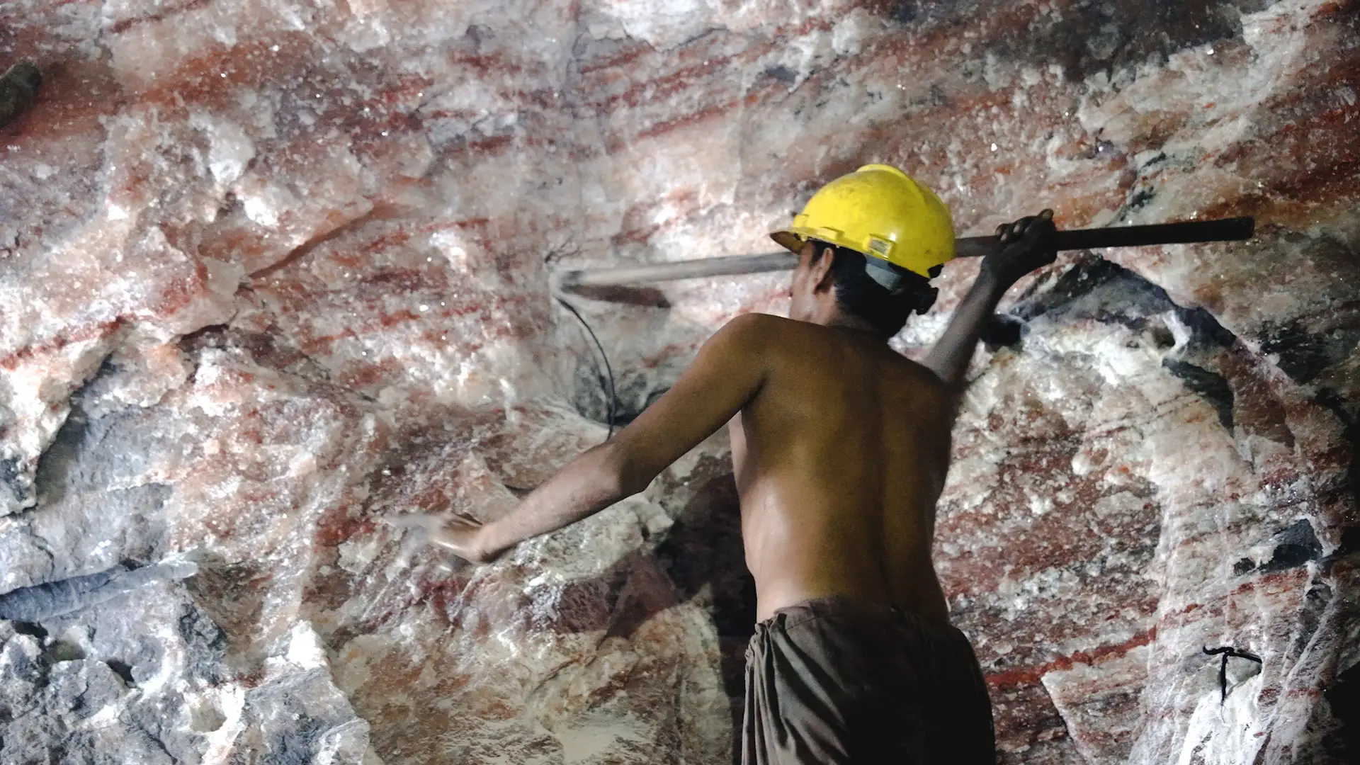 Worker in no shirt and yellow helmet packs explosives into the side of a salt mine.
