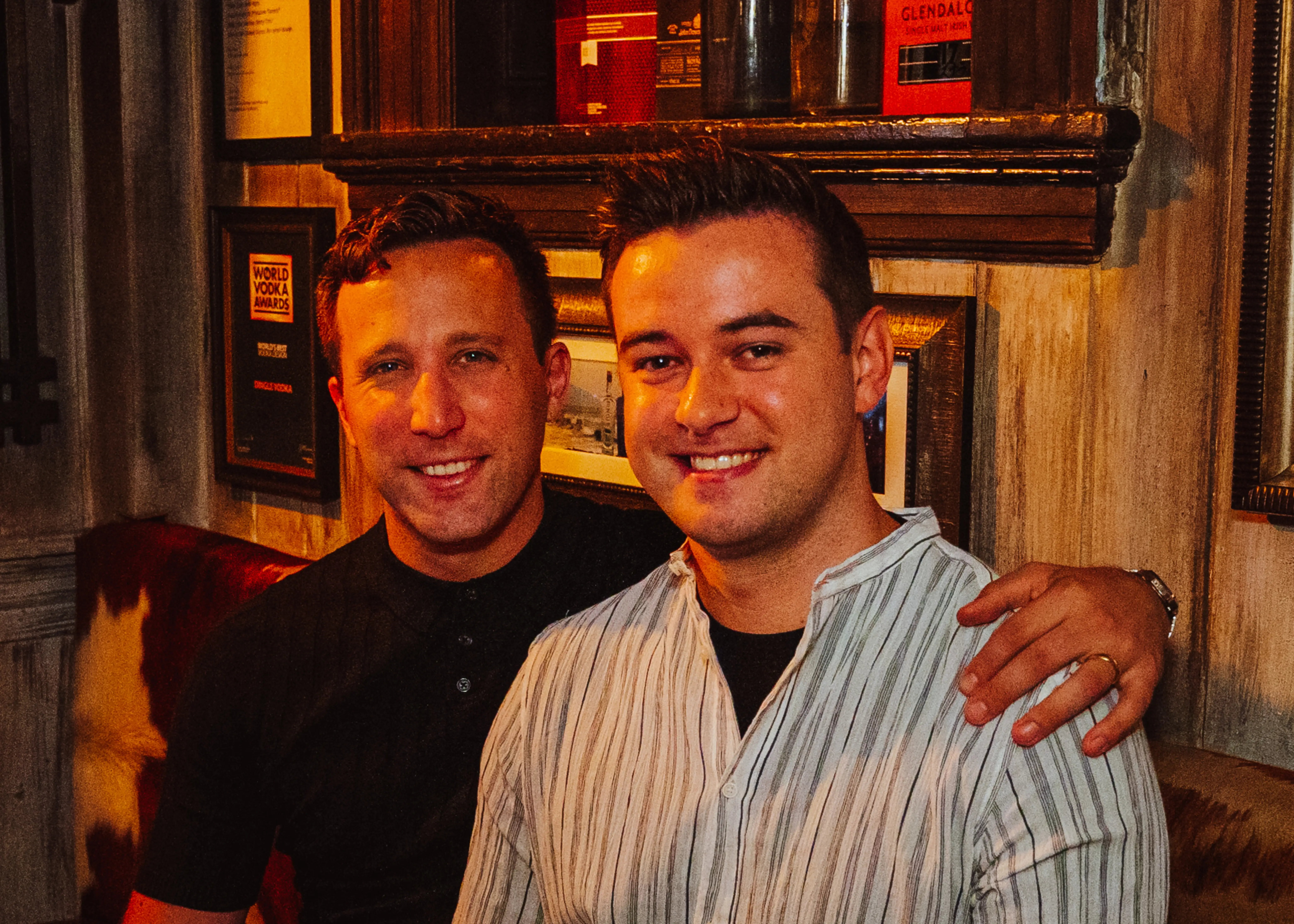Two men (Matt Cortland and John Fleming) in a dimly-lit room embrace in a side-hug.