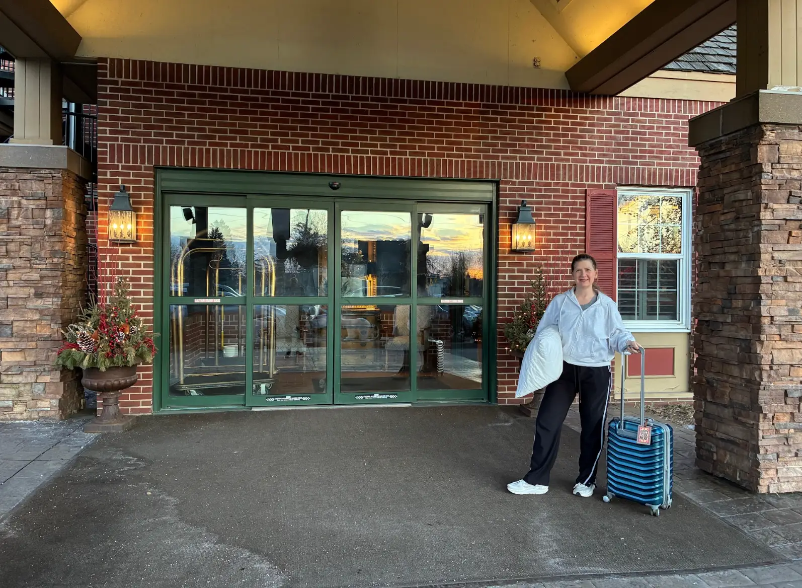 The writer holding a pillow and suitcase and standing in front of a hotel.